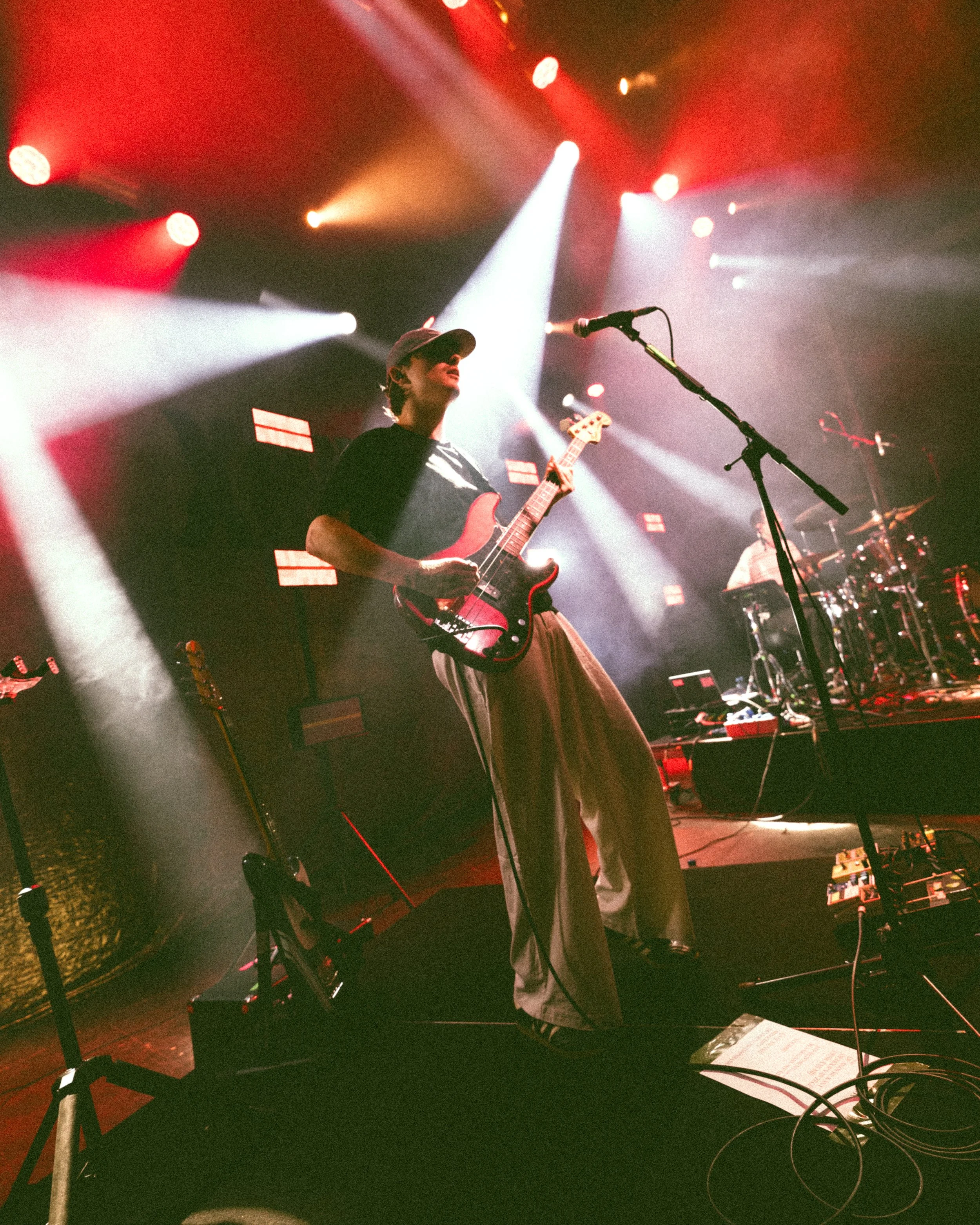 Musician playing electric guitar on stage with dramatic red and white stage lights, microphone stand in foreground, drums visible in background.