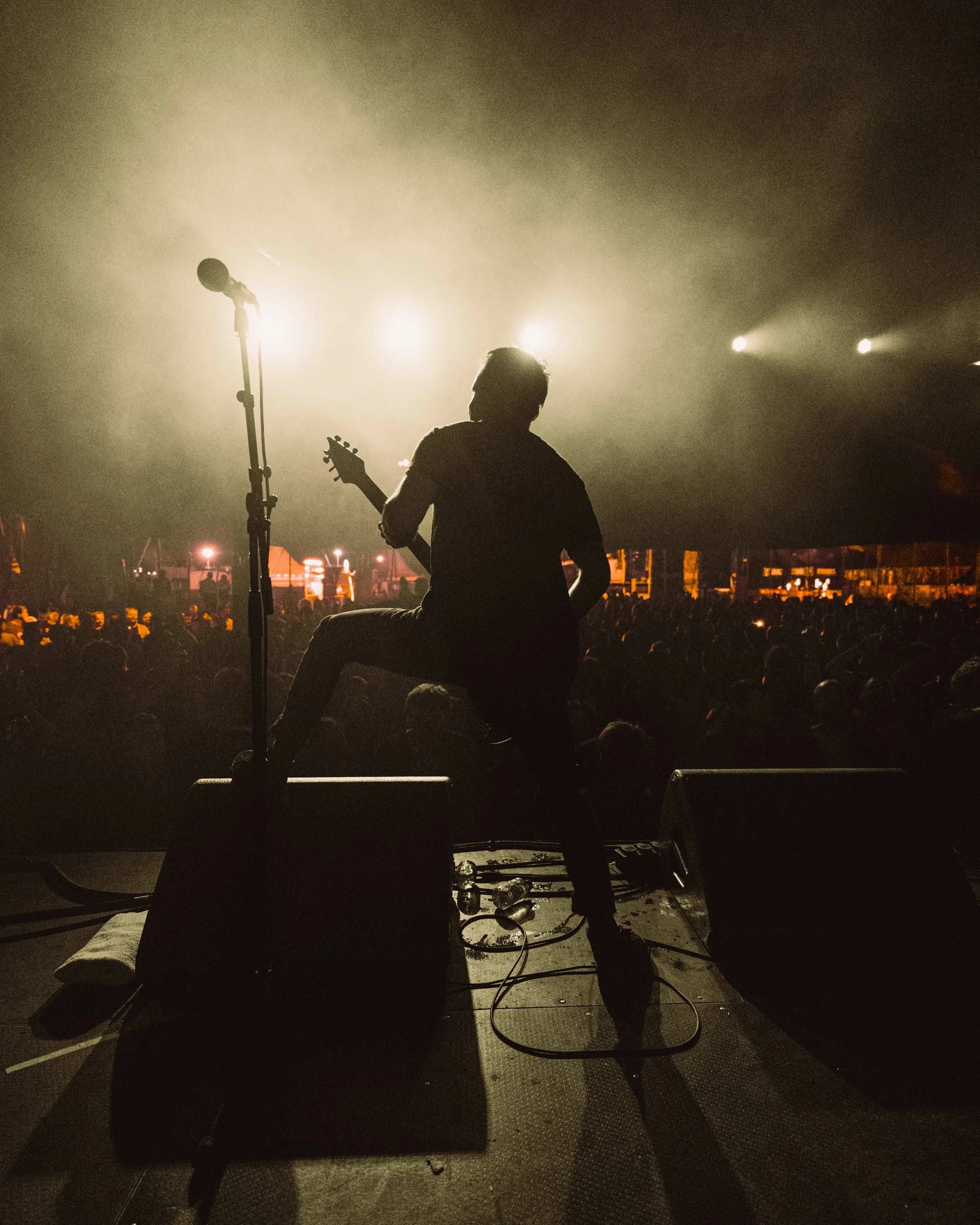 Silhouette of a musician playing guitar on stage at a concert with bright stage lights and a crowded audience in the background.