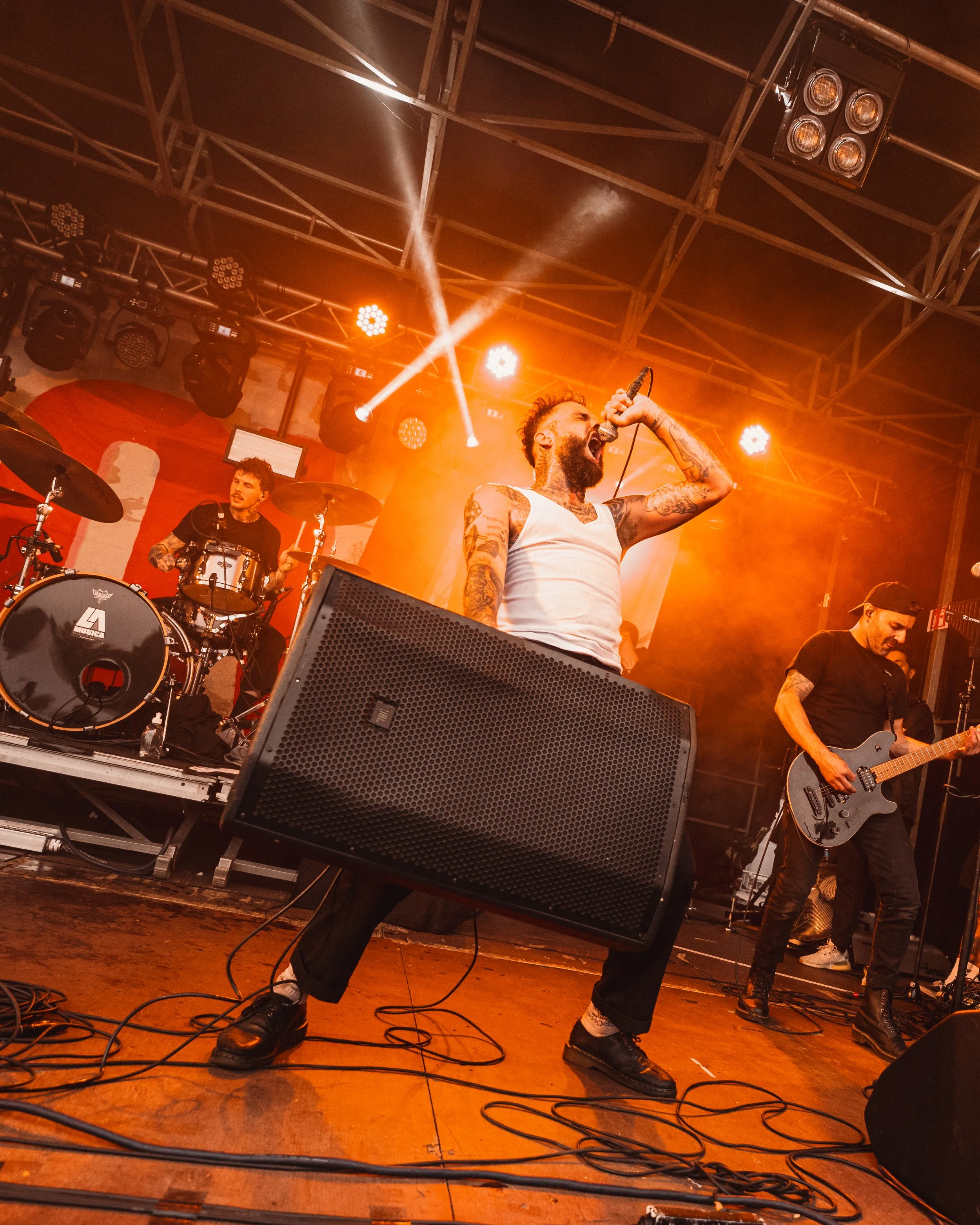 A band performs on a stage with orange lighting, including a vocalist with tattoos, a drummer, and a guitarist.