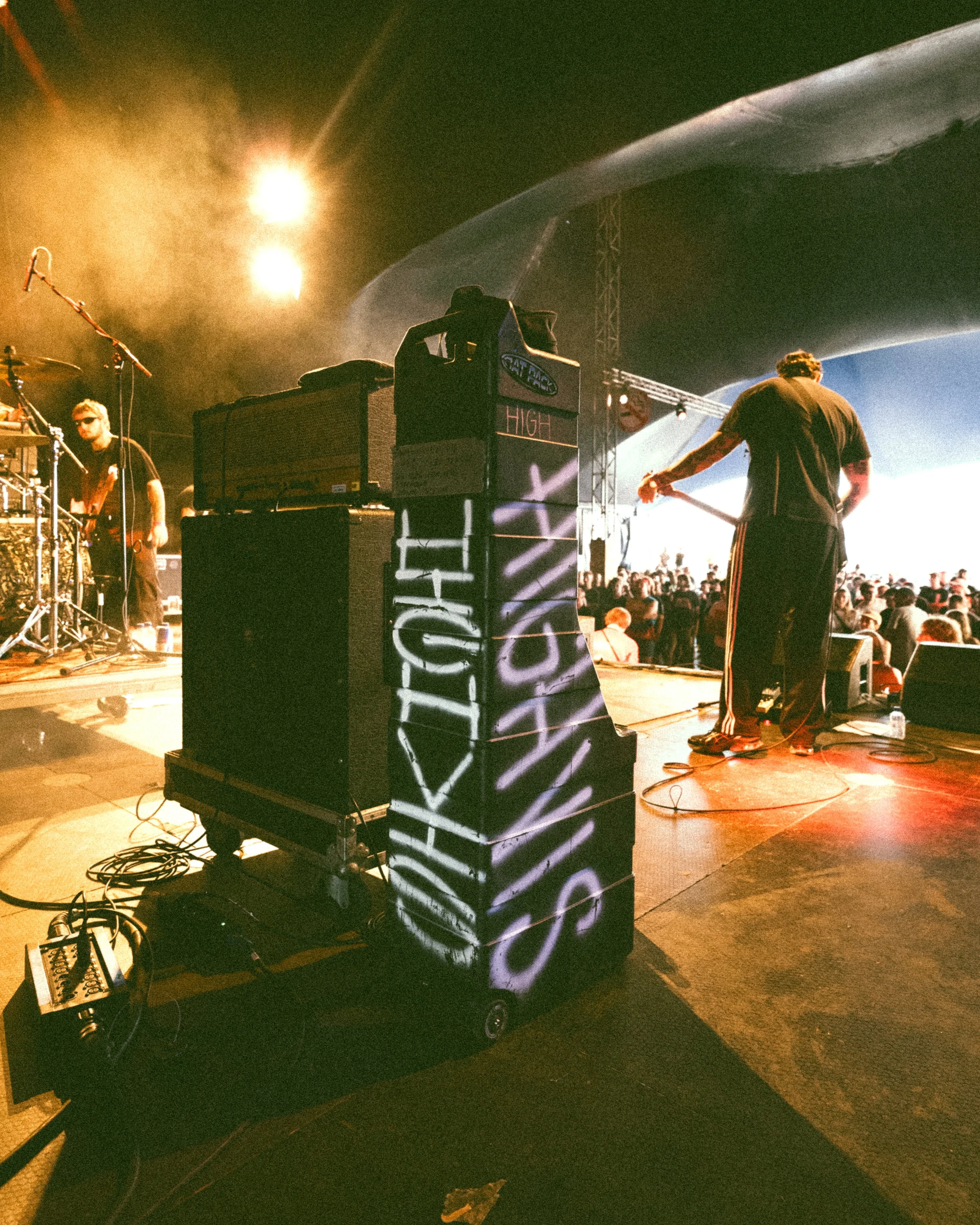 View of a live music performance on stage with two musicians, one playing drums and the other playing guitar, with stage lights and a crowd in the background. The stage has graffiti writing on a vertical surface.