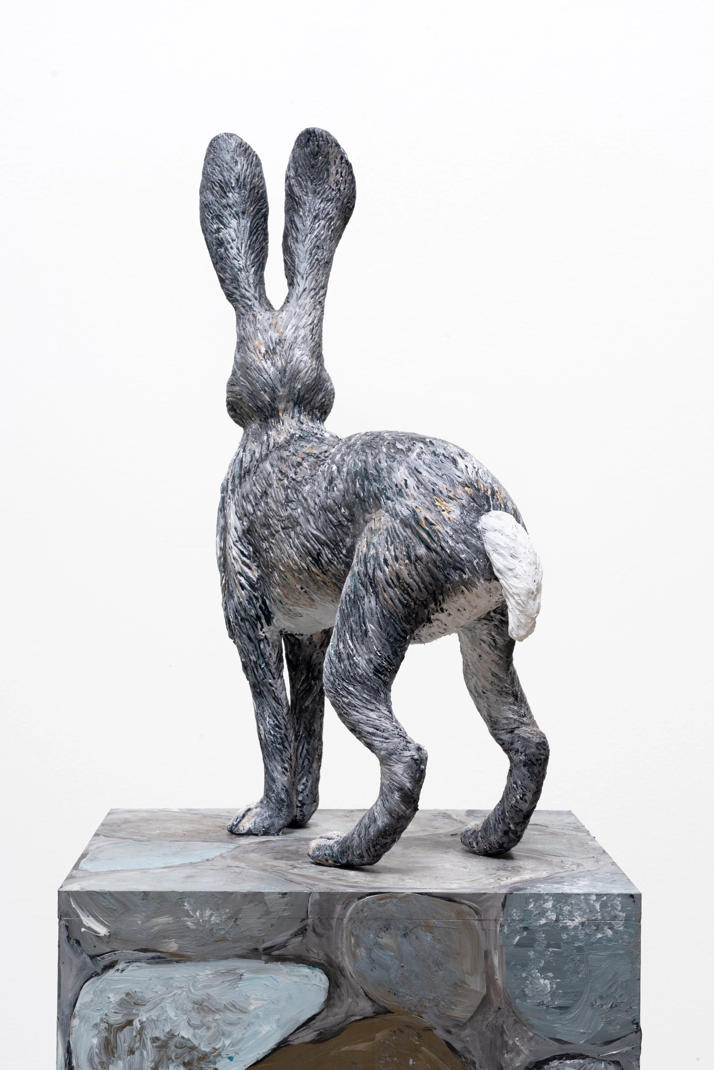 A sculpture of a rabbit with exaggerated features, painted in shades of gray, white, and black, placed on a painted pedestal against a plain white background.