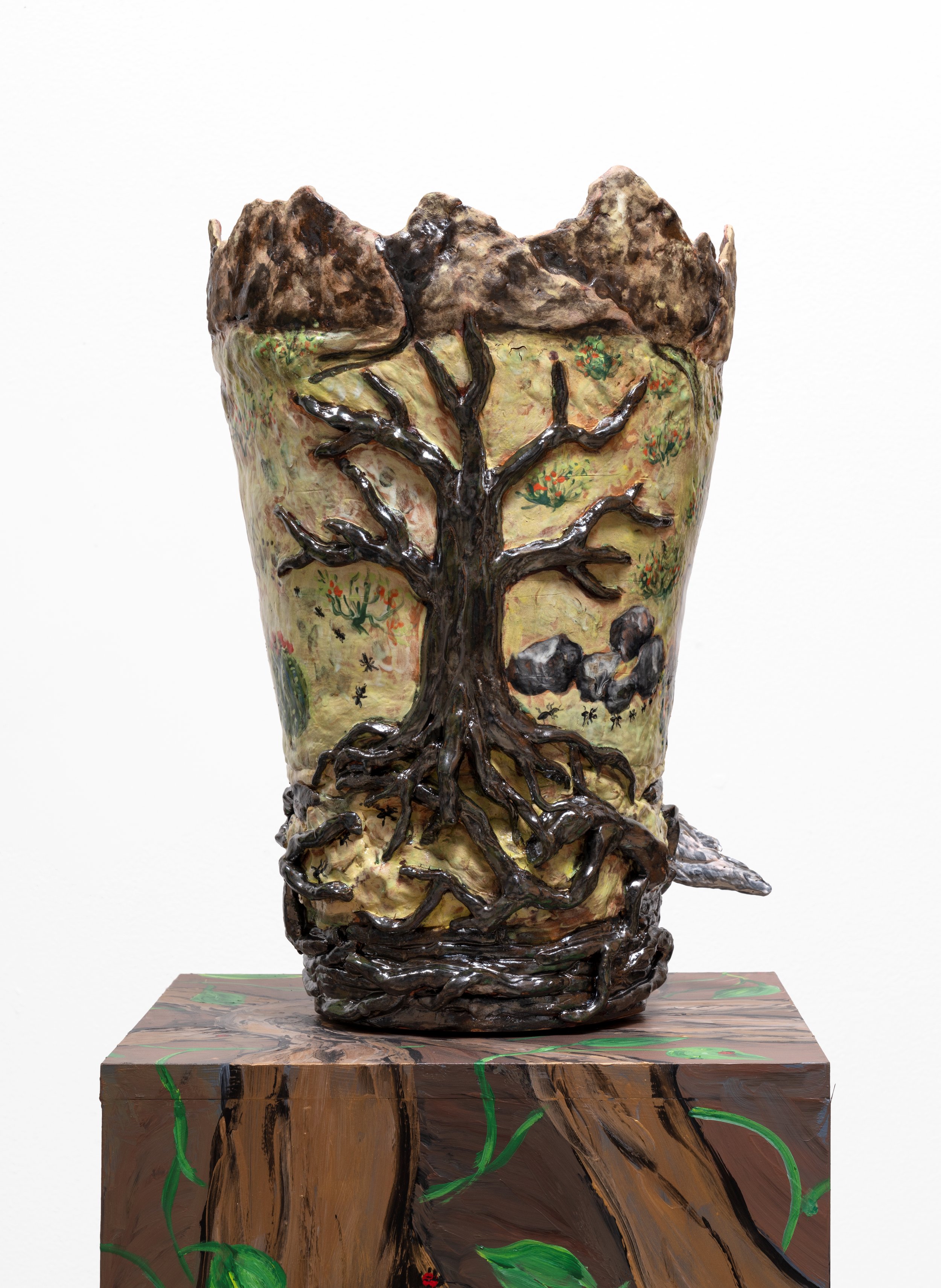 A ceramic or sculpture piece featuring a tree with dark, textured branches and roots, painted on a yellowish background with small painted flowers, positioned on a wooden base with green accents.