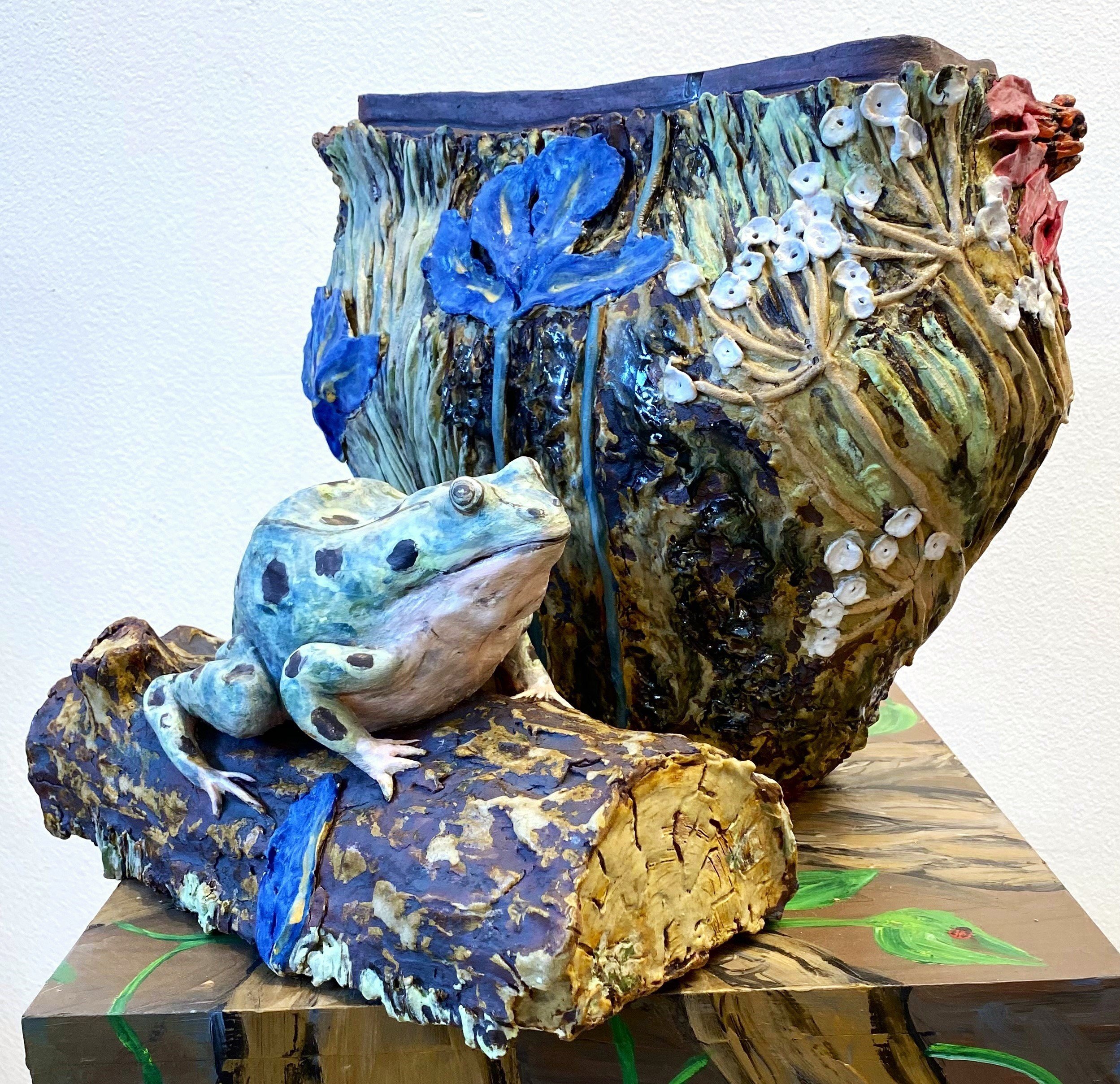 Ceramic art sculpture of a frog sitting on a log with large ornate vase or pot in the background, featuring hand-painted floral and nature designs, displayed on a painted wooden surface.