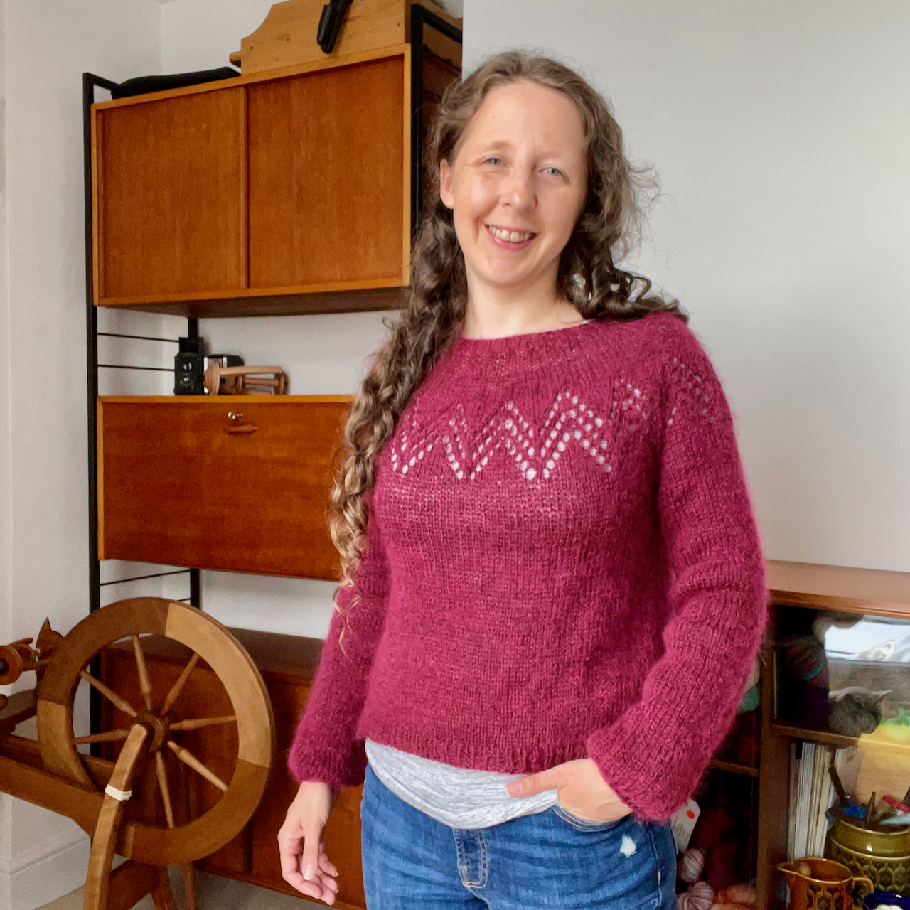 Becks, a white woman with long, light brown curly hair, wearing a slightly cropped handknit raspberry pink sweater with a chevron lace yoke and long balloon sleeves