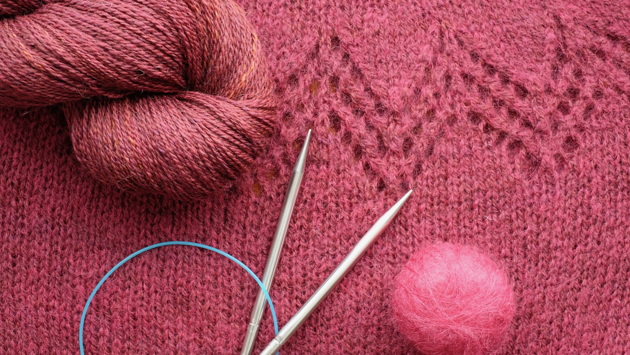 A hank of burgundy handspun yarn on top of a piece of knitted lace fabric, alongside a pair of knitting needles and a small ball of mohair.