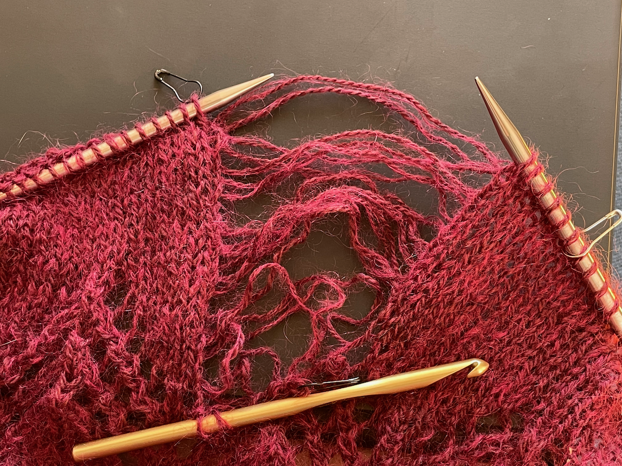 Knitting needles hold most of the stitches but a large chunk have been dropped back, leaving strands of yarn floating around, the only active stitches being held by a crochet hook.