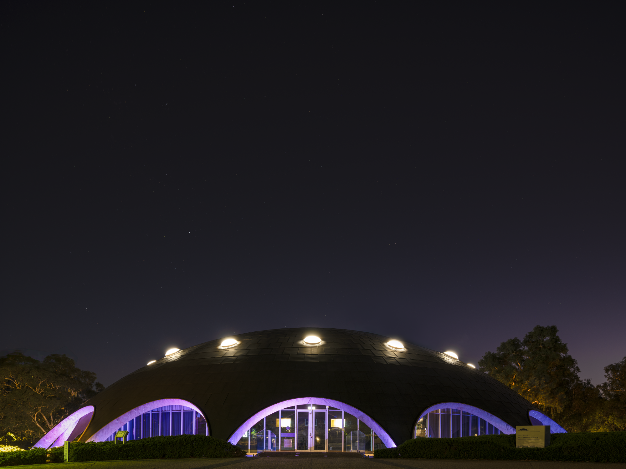 A dome-shaped building at night illuminated with purple and white lights, with trees and a dark starry sky in the background.