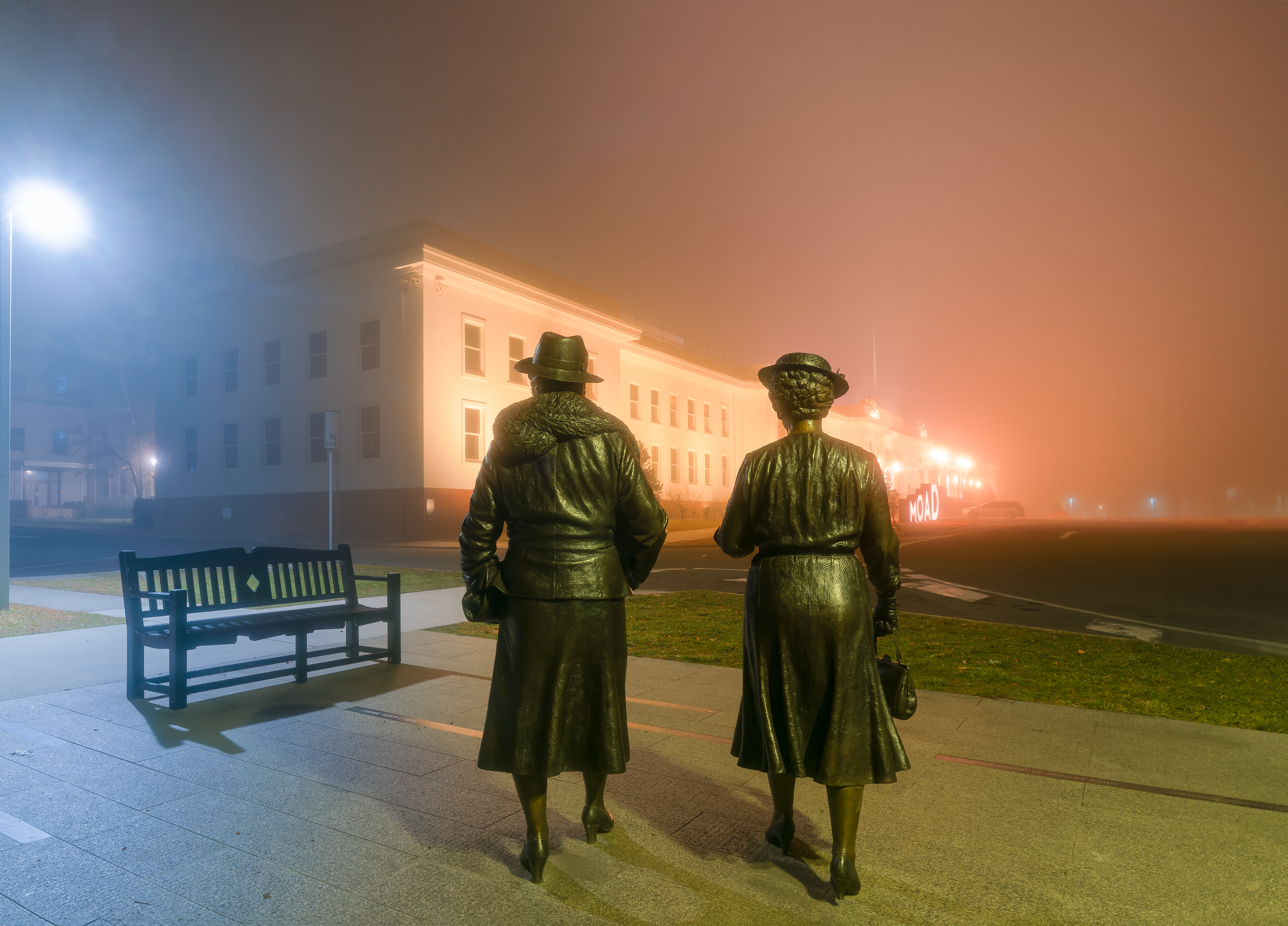 Bronze statues of a man and woman walking together in a foggy street scene at night, with a lit-up building and neon sign in the background.