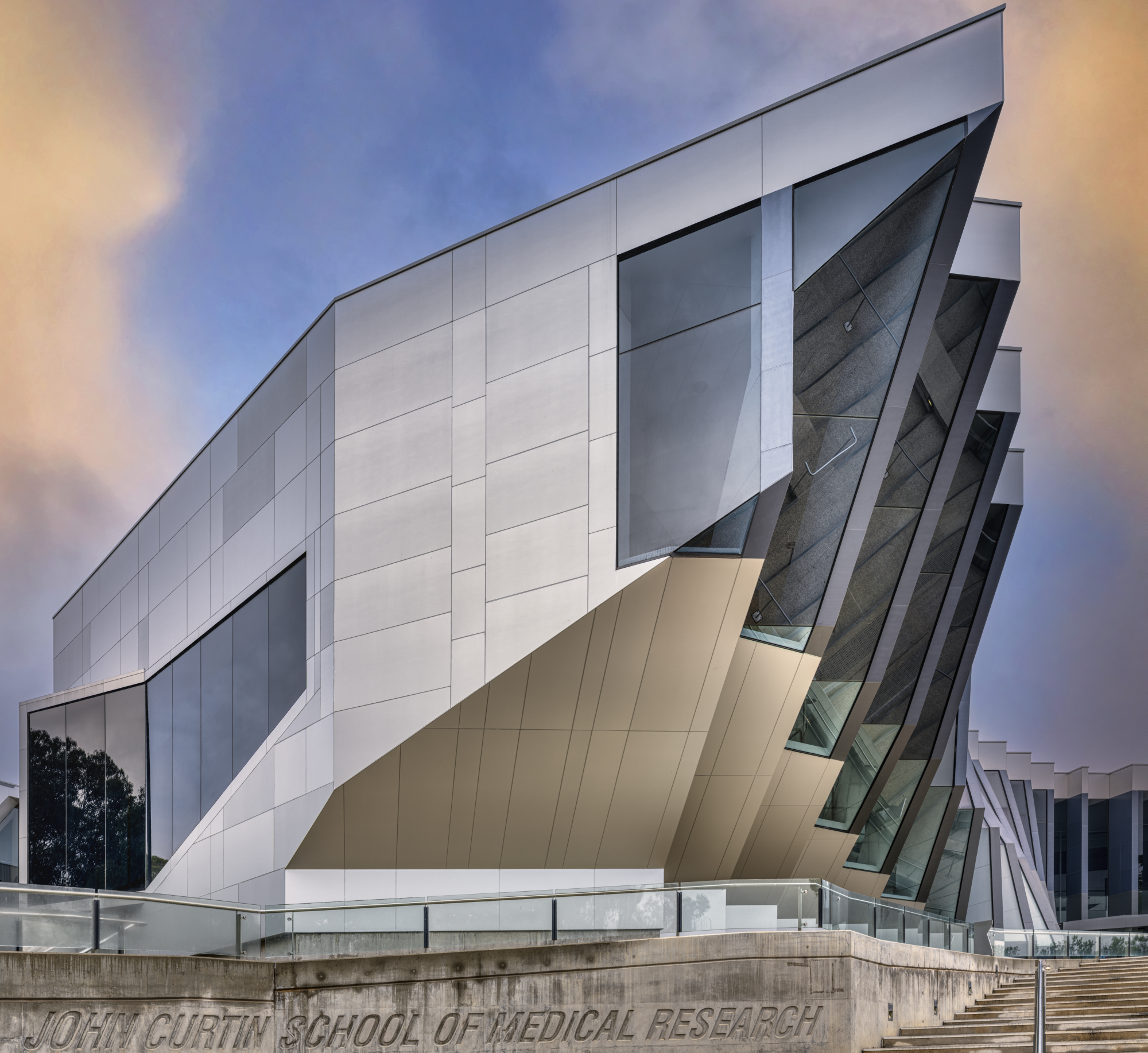 Modern building with angular architecture and large glass windows, part of the John Curtin School of Medical Research, against a colorful sky.