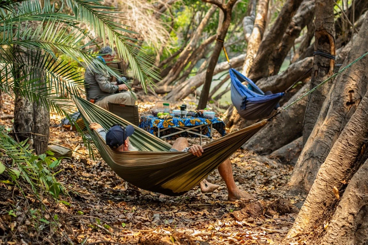 Two men relaxing in hammocks in a wooded area with trees and fallen leaves, with a table set with snacks and drinks between them.
