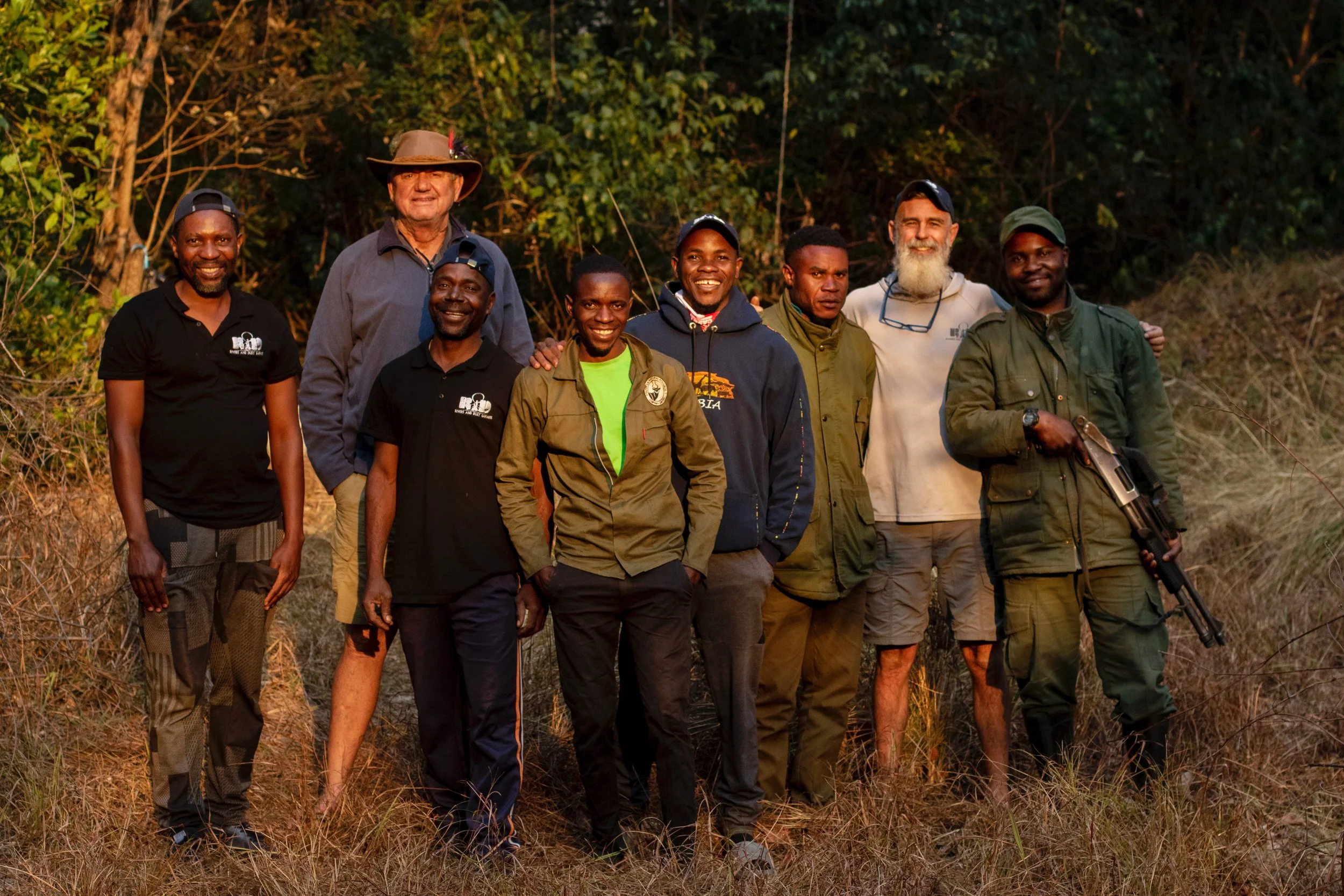 Group of eight men standing outdoors in a natural setting with trees and dry grass, smiling at the camera, some wearing outdoor and hunting gear.