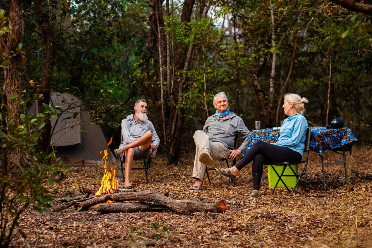 Three people sitting around a campfire in a forest, enjoying a conversation, with a table nearby holding drinks and gear, and a tent in the background.