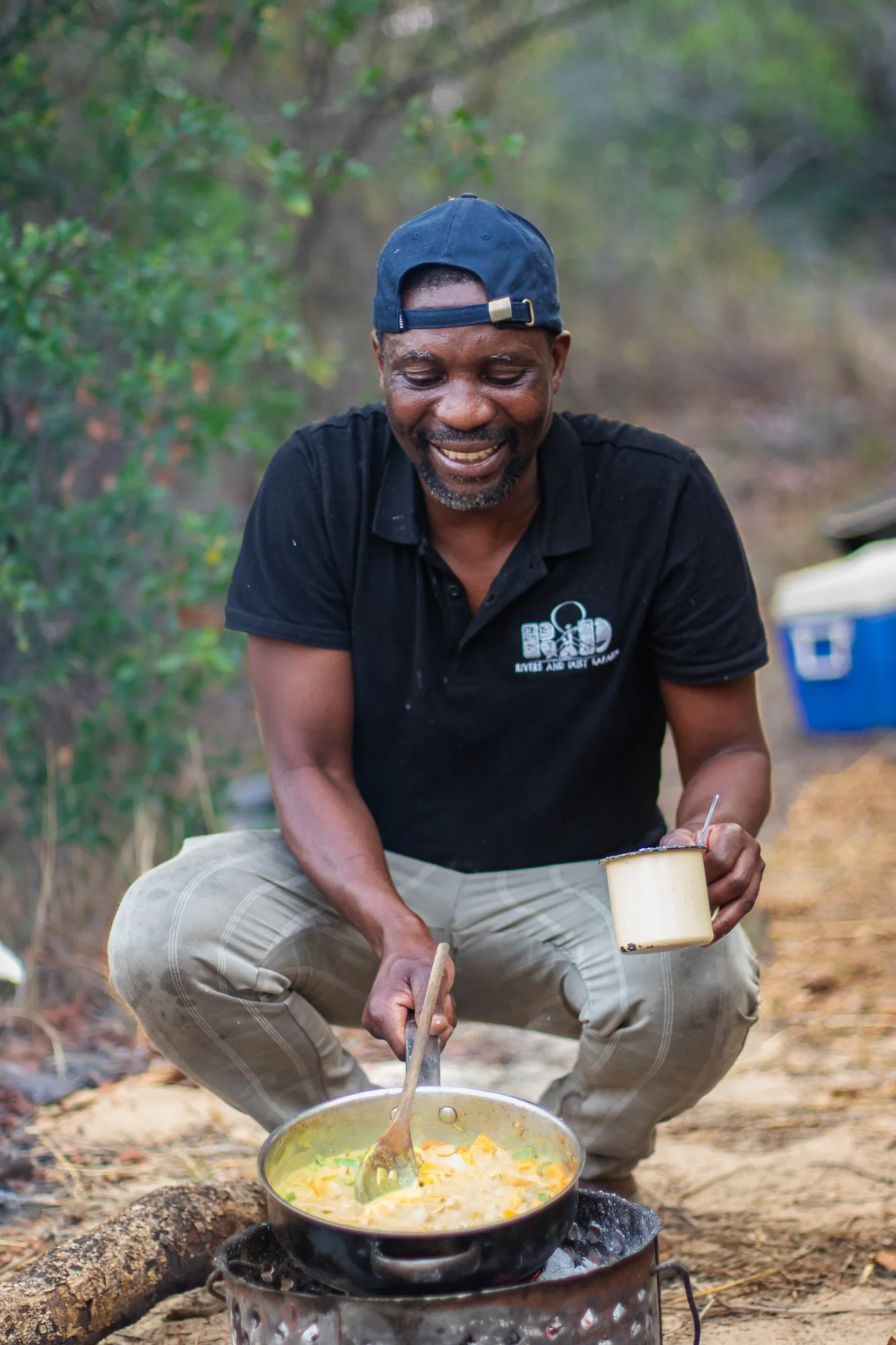 Man cooking food outdoors over a small fire in a forest setting. He is smiling and holding a spoon and a cup, wearing a black shirt and a backwards cap.
