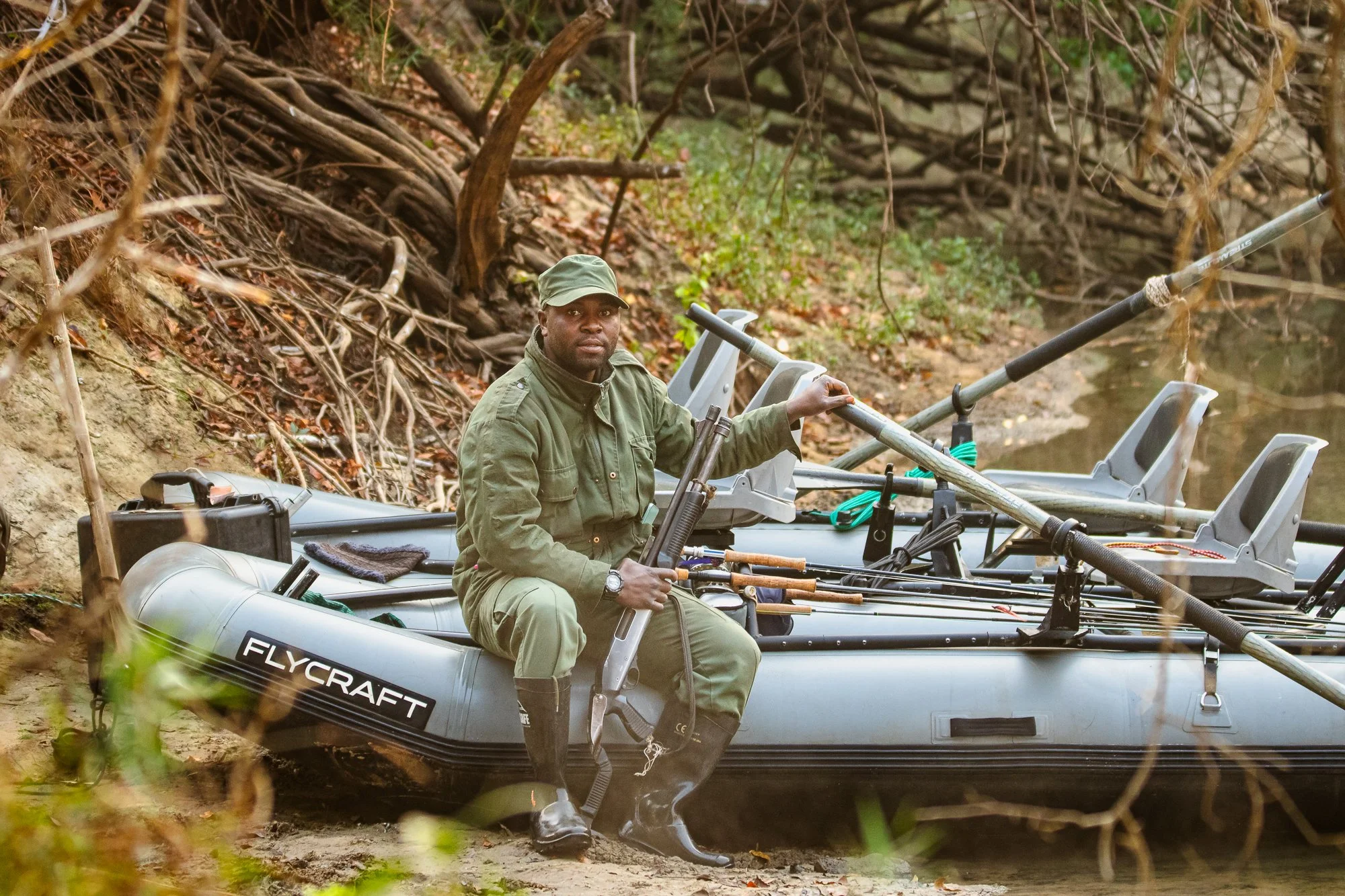 A person in green outdoor gear sitting in a boat labeled 'Flycraft' on a riverbank, holding photography or videography equipment.