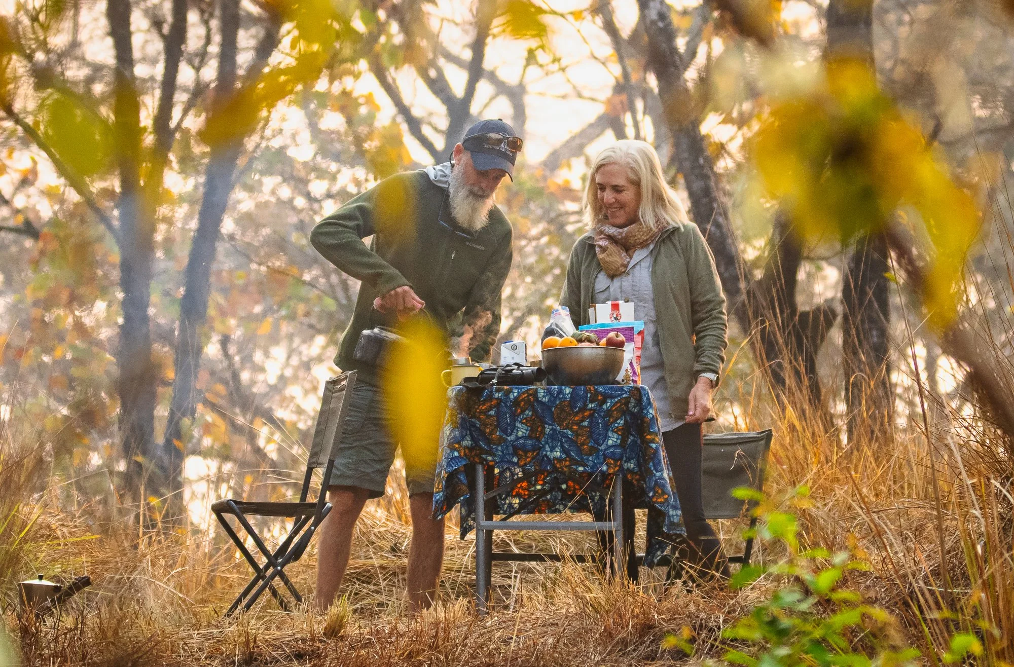 A man and a woman enjoying a meal outdoors in a wooded area during autumn, with a table covered with a colorful cloth, holding a bowl of fruit and other food items, surrounded by camping chairs.