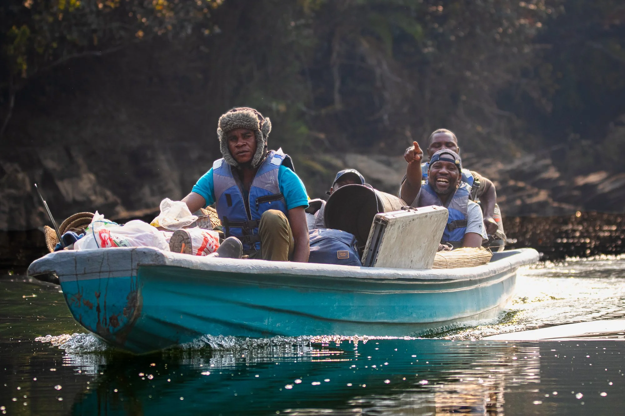 A group of men on a small boat filled with supplies, wearing life jackets, traveling on a river surrounded by rocky terrain and trees.