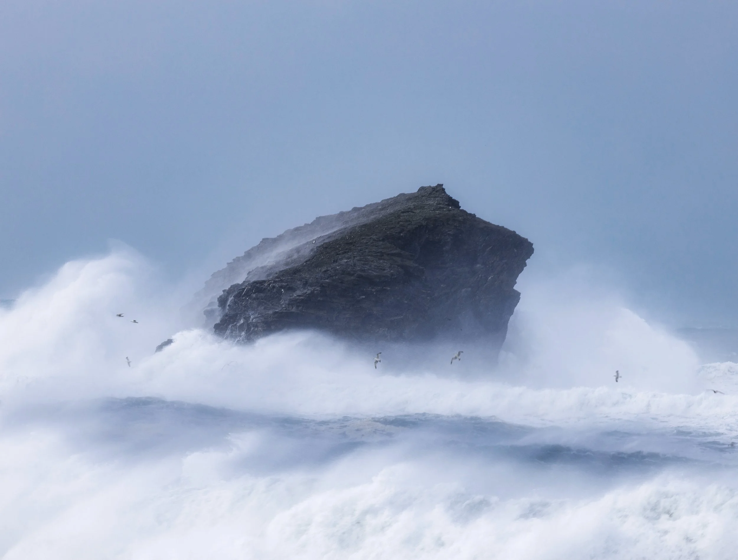 Large crashing waves hit a rocky outcrop in the ocean, with seagulls flying above.
