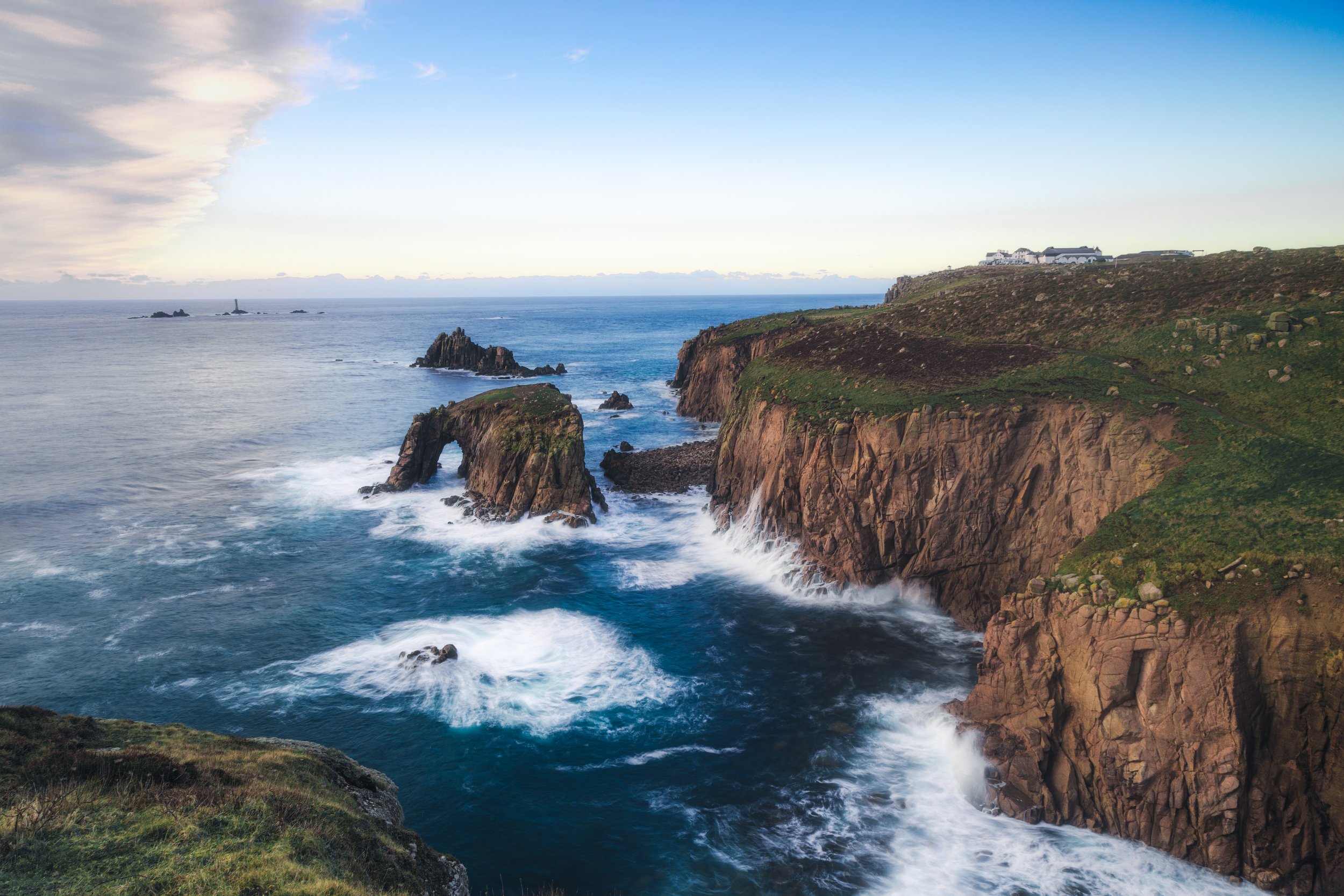 Cliffs along a rugged coastline with green grass, rocky formations, and waves crashing against the rocks, under a partly cloudy sky.