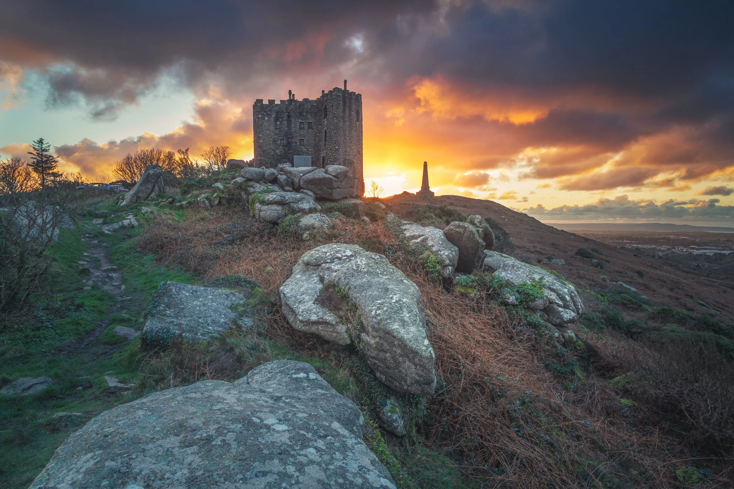 A castle on a hill during sunset, with rocky terrain and a trail leading up to it, and a monument in the background.