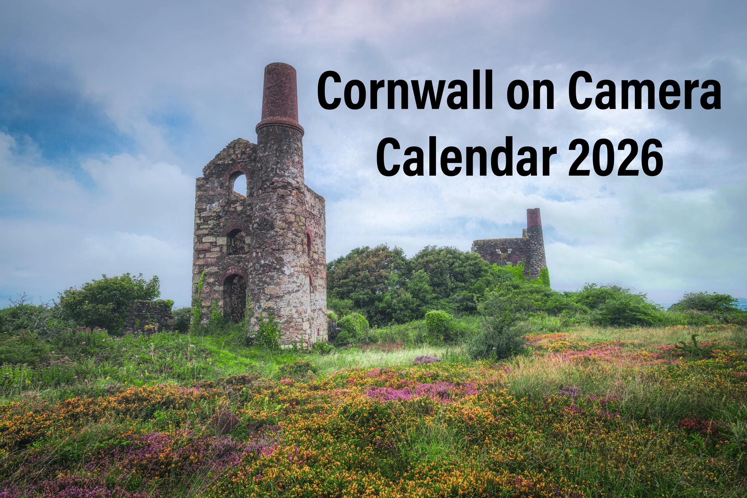 Ruined stone structures, one with a tall chimney, surrounded by green vegetation and colorful wildflowers under a partly cloudy sky, with black text overlay reading 'Cornwall on Camera Calendar 2026'.