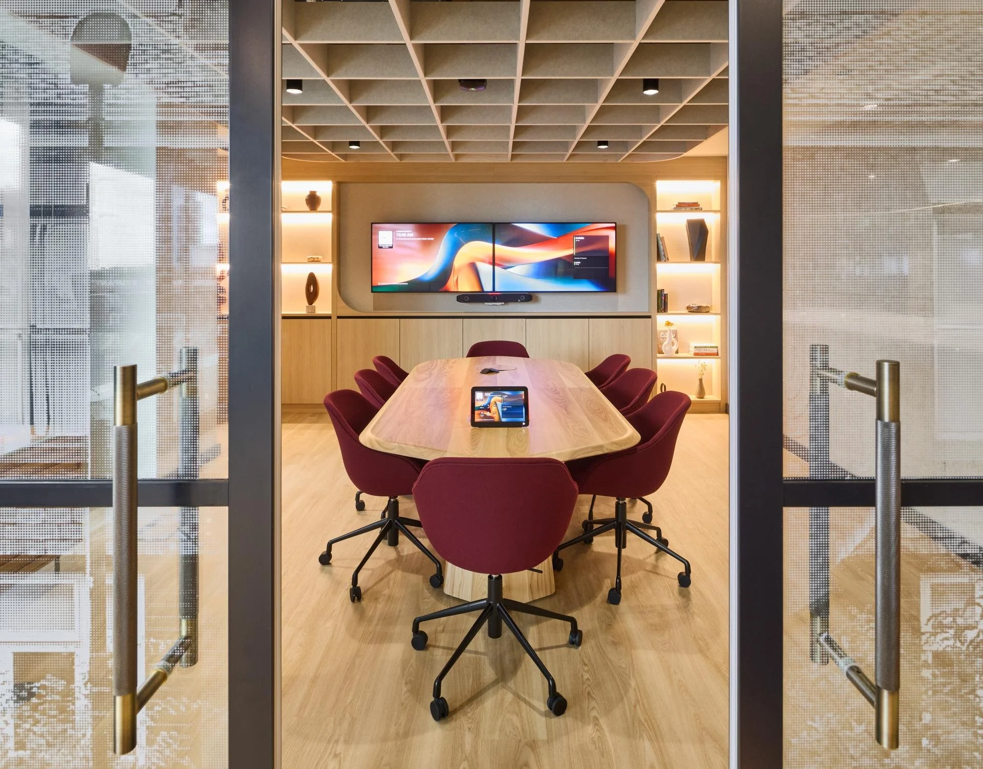 A modern conference room with a large wooden table surrounded by six burgundy office chairs. There are glass sliding doors with gold handles. In the background, a large screen is mounted on a beige wall, with a soundbar below it. The room has built-in shelves with decorative items and books, illuminated with warm lighting.