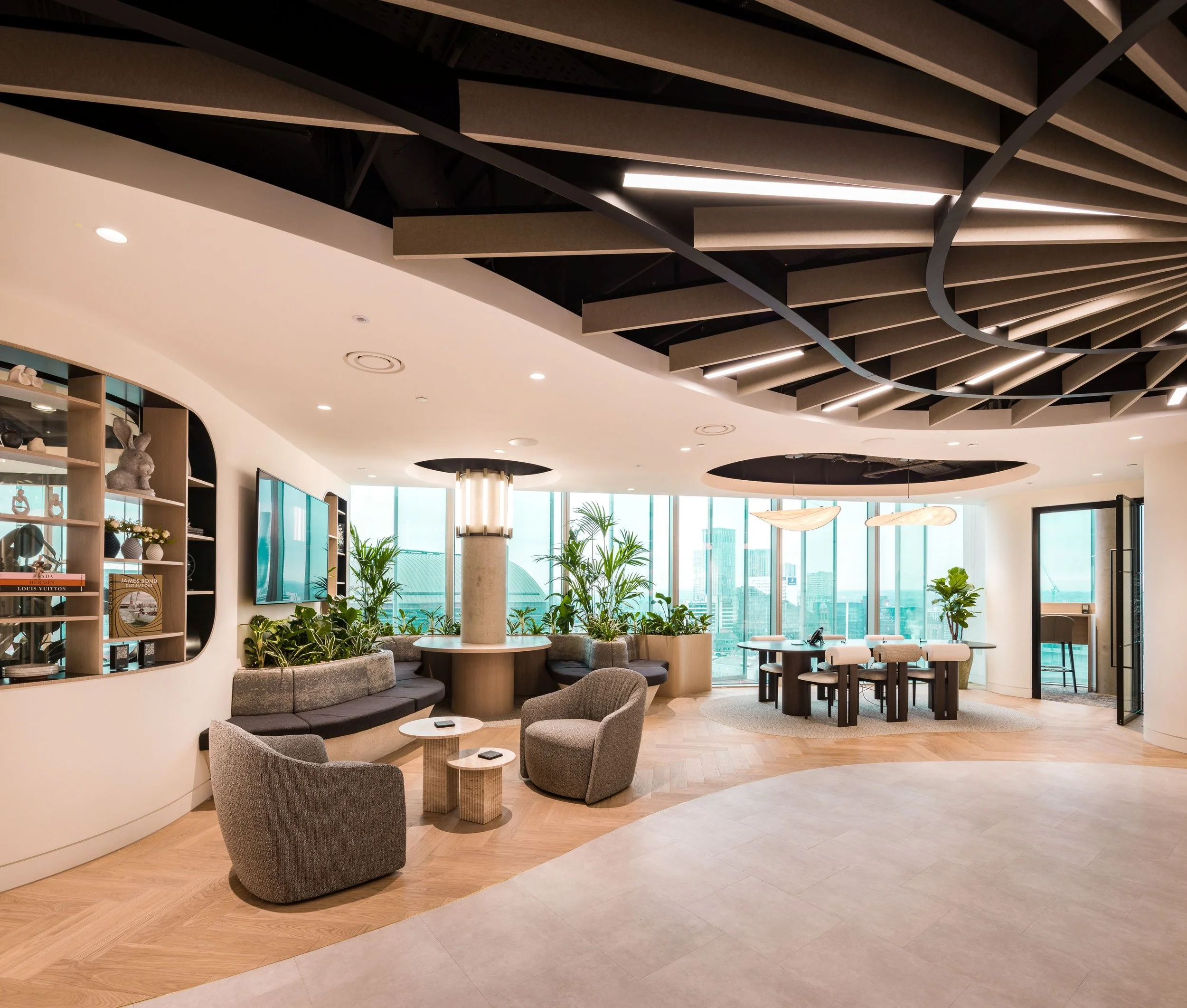 Modern office lounge with large windows, comfortable seating, plants, and contemporary decor.