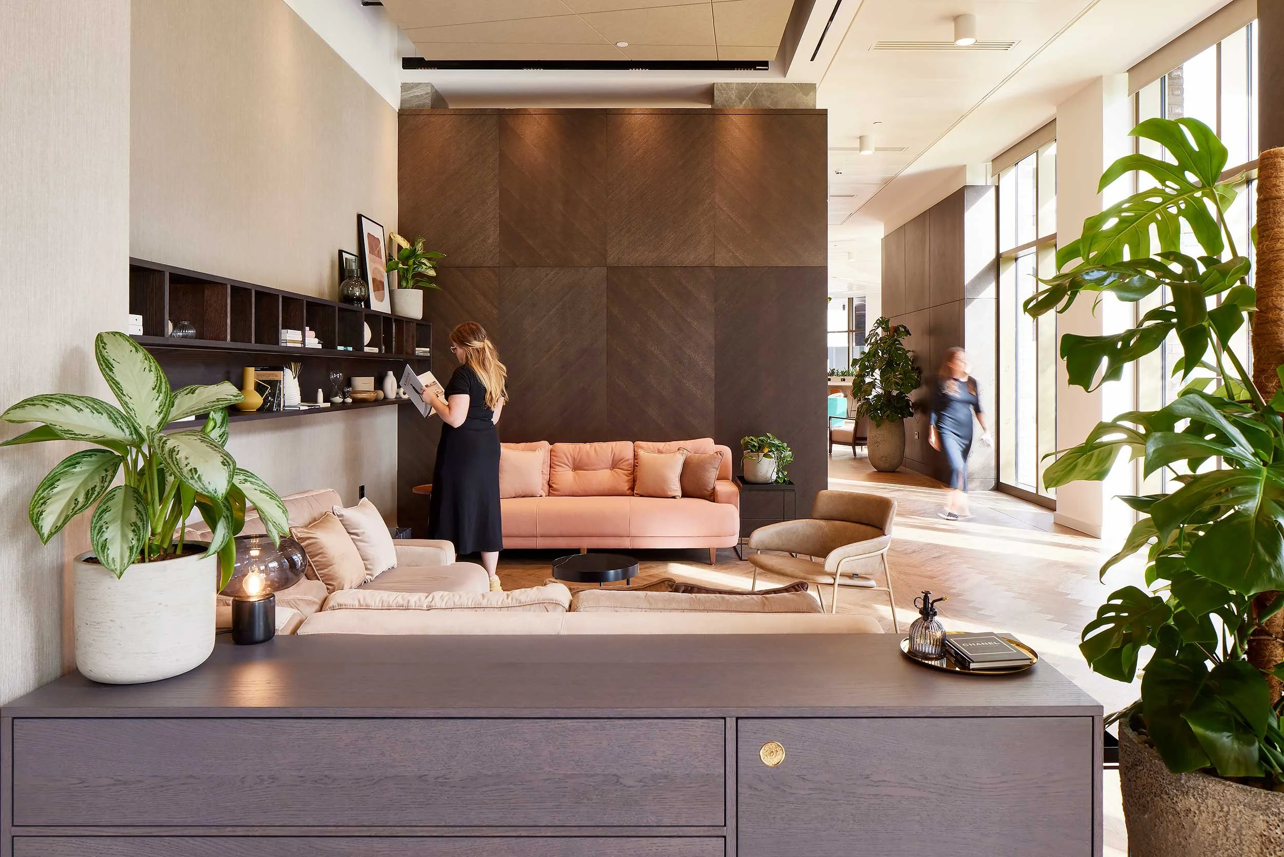 Modern hotel lobby with a woman reading at a shelf, pink and beige sofas, and large windows with natural light.