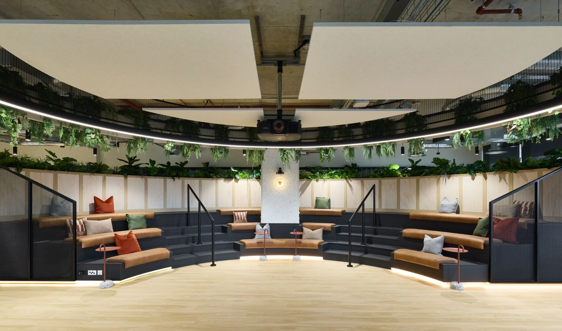 A modern indoor seating area with curved tiered black seating, beige cushions, and colorful throw pillows. There are plants and greenery along the back wall and hanging pot plants above. The ceiling has large white panels, a projector, and industrial elements. The floor is light wood, and subtle LED lighting accents the edges of the seating area.