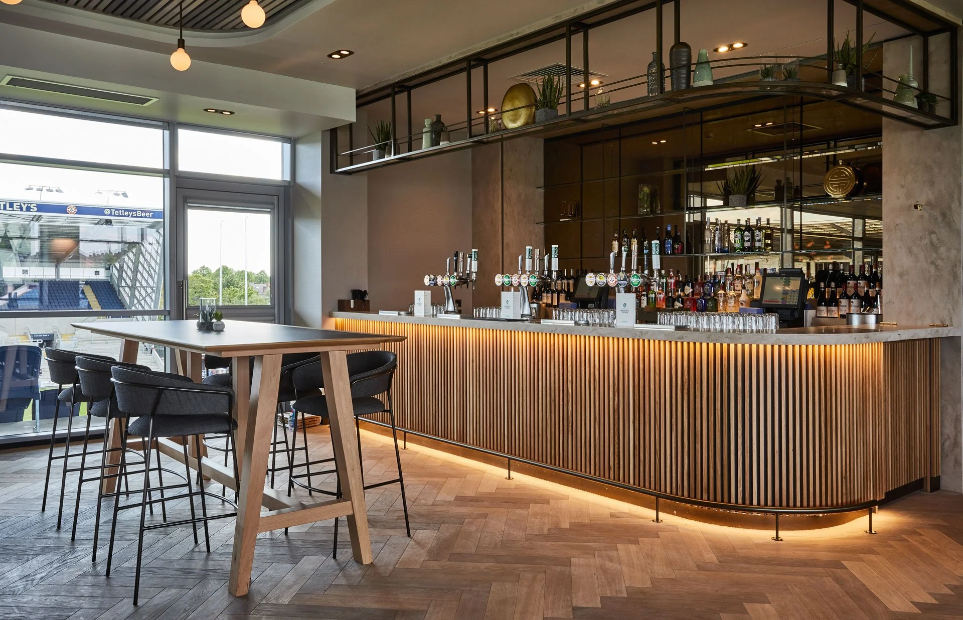 Modern bar with wooden facade and liquor bottles on shelves, large window revealing stadium outside, high table with black chairs, and overhead lighting.