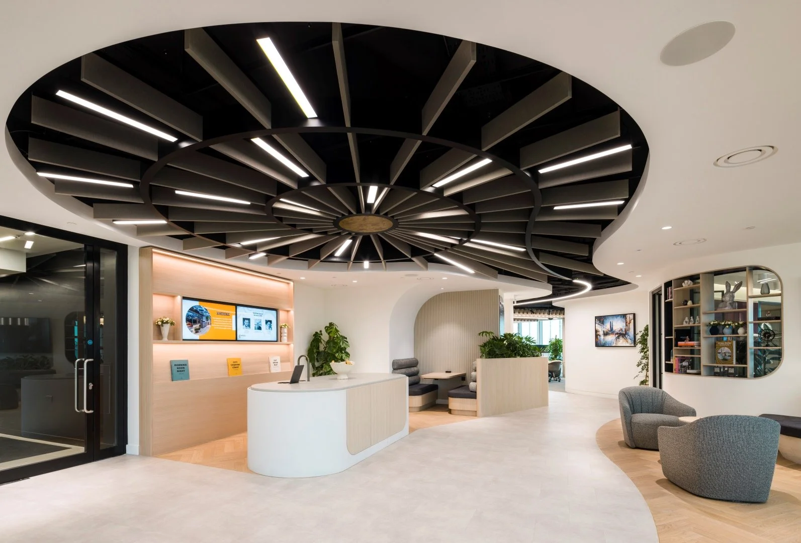 Modern hotel lobby with a circular white reception desk, black geometric ceiling structure with integrated lighting, gray and white seating area, potted plants, a bookshelf, and a wall-mounted television.