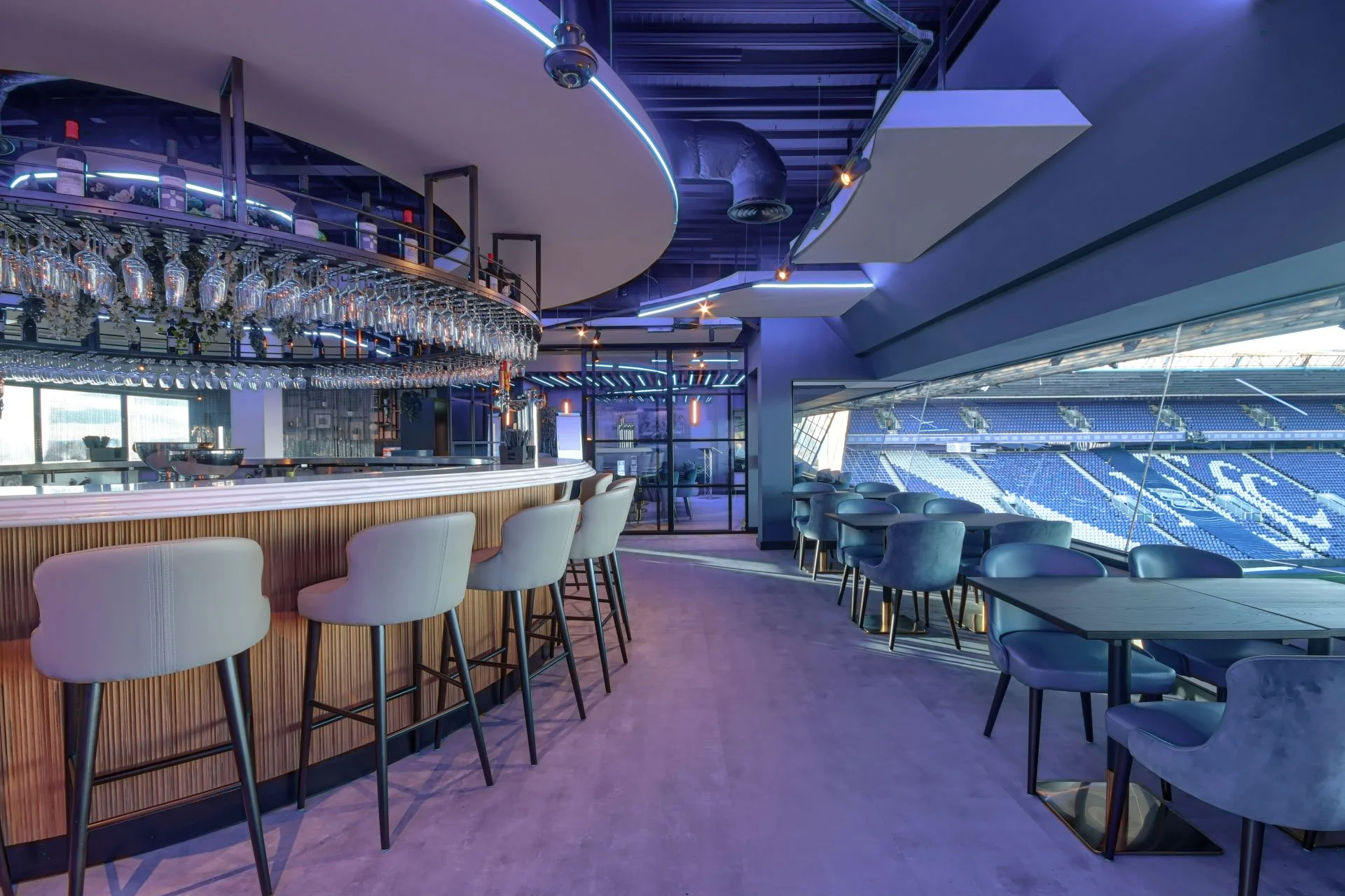 Interior of a modern sports stadium lounge with a bar area featuring hanging wine glasses, seating with plush chairs, large windows overlooking the empty stadium seats, and blue-purple ambient lighting.