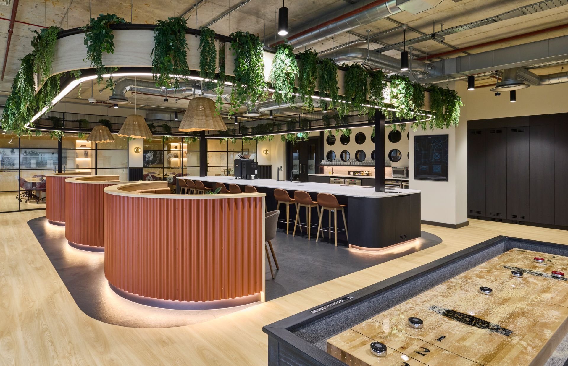 Interior of a modern recreational space featuring a bar with high chairs, hanging pendant lights, green plants, and an ice hockey table in the foreground.