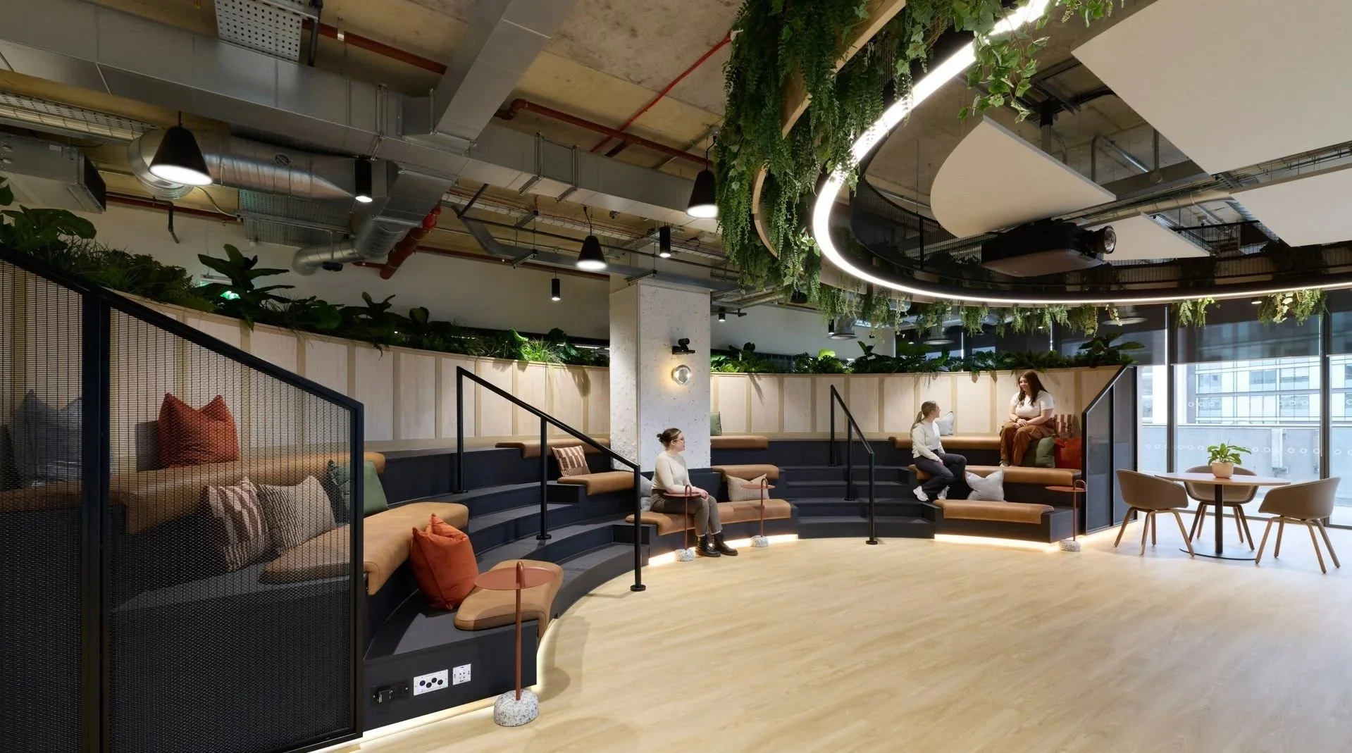 Modern indoor lounge with tiered seating, cushions, and potted plants, featuring curved design and large windows providing natural light.