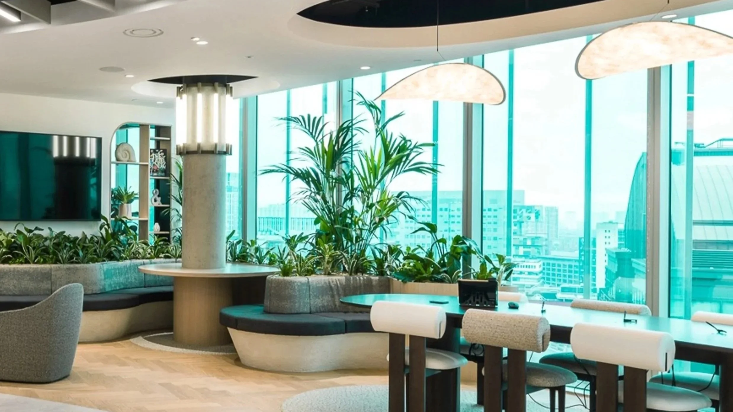 Curved breakout seating and shared worktable set beside floor-to-ceiling glazing with city views, featuring integrated planting, soft upholstered furniture and sculptural lighting in a contemporary commercial workplace interior.