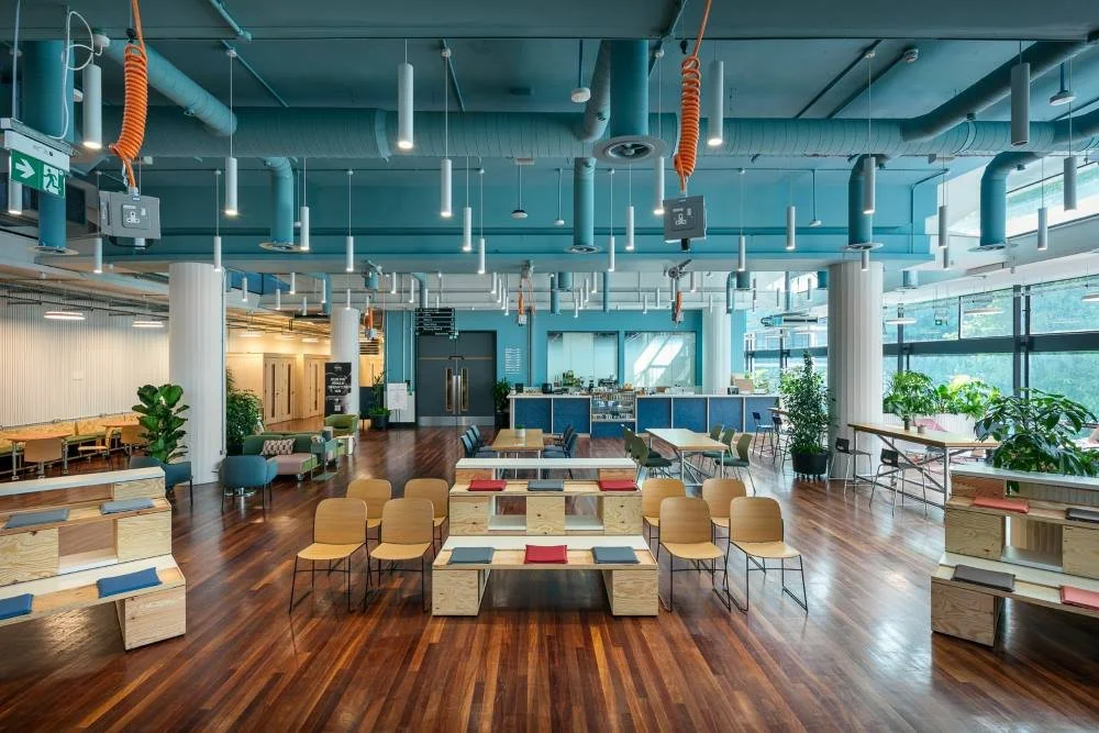 A spacious indoor area with modern decor, wooden floors, green plants, seating arrangements, and a coffee counter, likely a lounge or communal space in an office or public building.