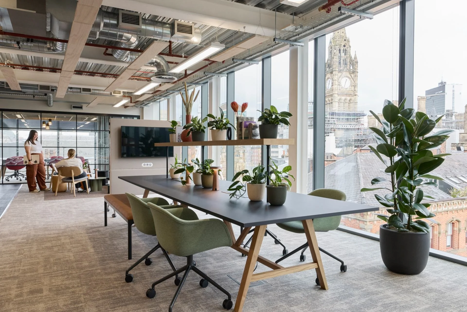 Modern office with large windows, potted plants, a conference table with green chairs, and cityscape view including a clock tower.