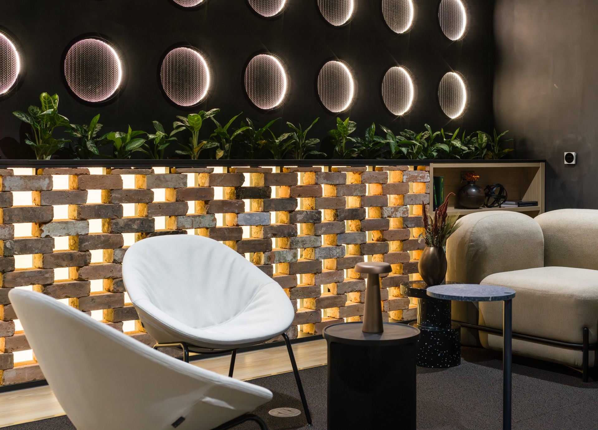 Modern living room with a brick wall partition, indoor plants, circular wall art, and stylish furniture including a white lounge chair, beige sofa, and side tables with decorative vases.