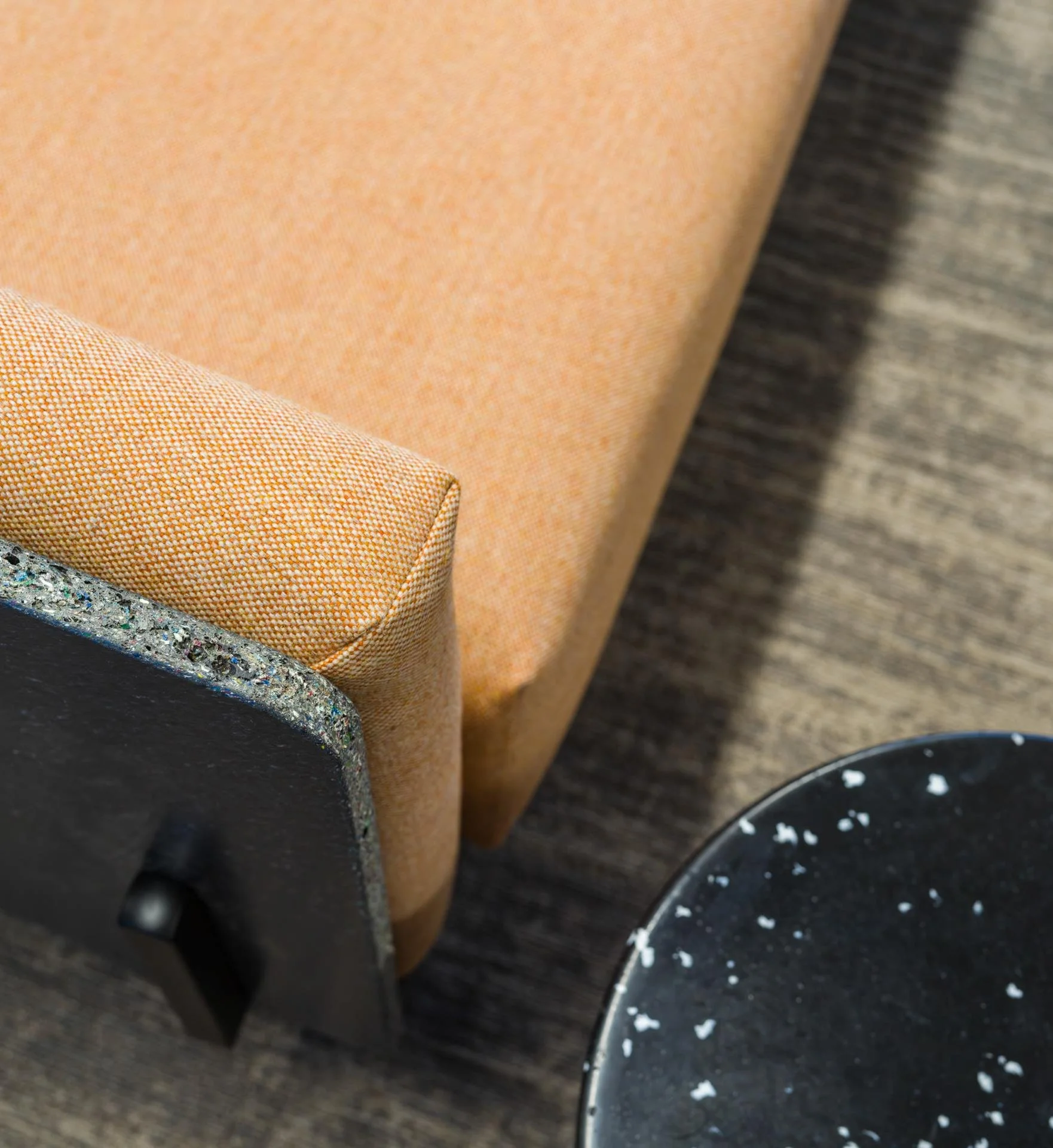 Close-up of a beige upholstered chair, black round table with white speckles, and gray carpeted floor.