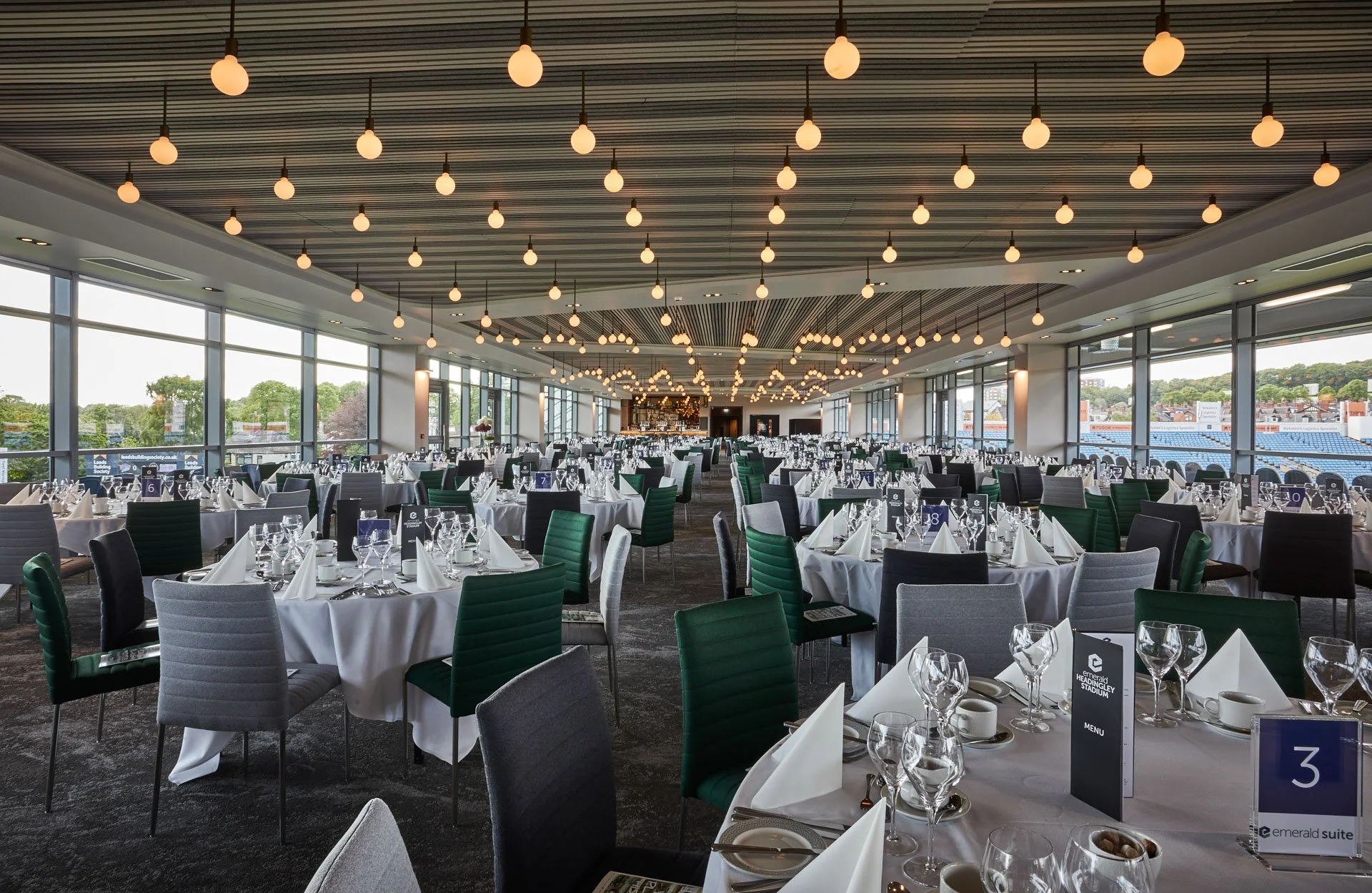 A large banquet hall set up for a formal event with round tables covered in white tablecloths, decorated with glassware, plates, and napkins, and surrounded by chairs in green, gray, and black. The hall has floor-to-ceiling windows on both sides, providing a view of trees and buildings outside. The ceiling features numerous hanging light fixtures, and the lighting is bright and modern.