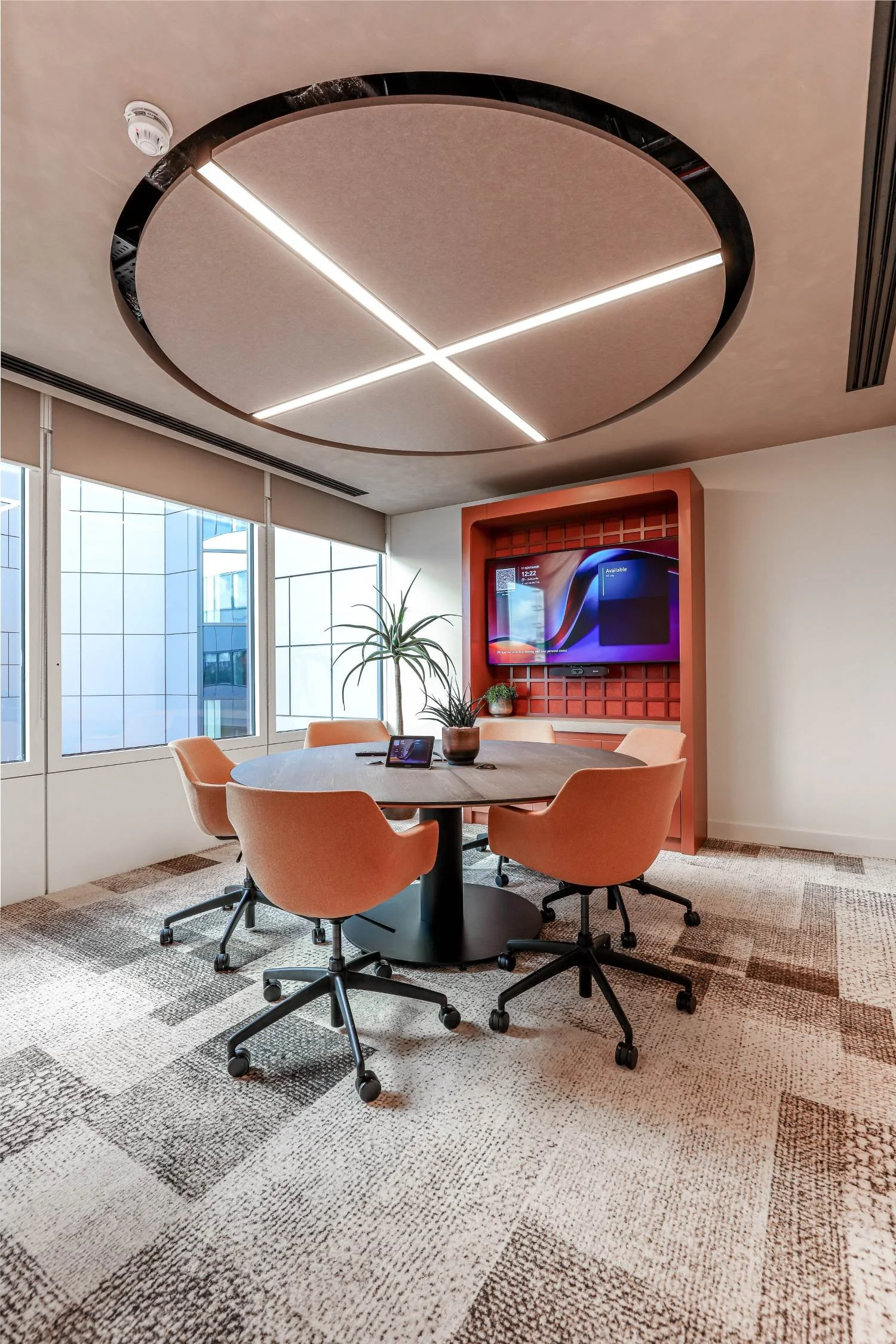 Modern conference room with round wooden table, six salmon-colored swivel chairs, a large flat-screen TV mounted on an orange feature wall, a potted plant, a tablet on the table, and large windows with blinds letting in natural light.