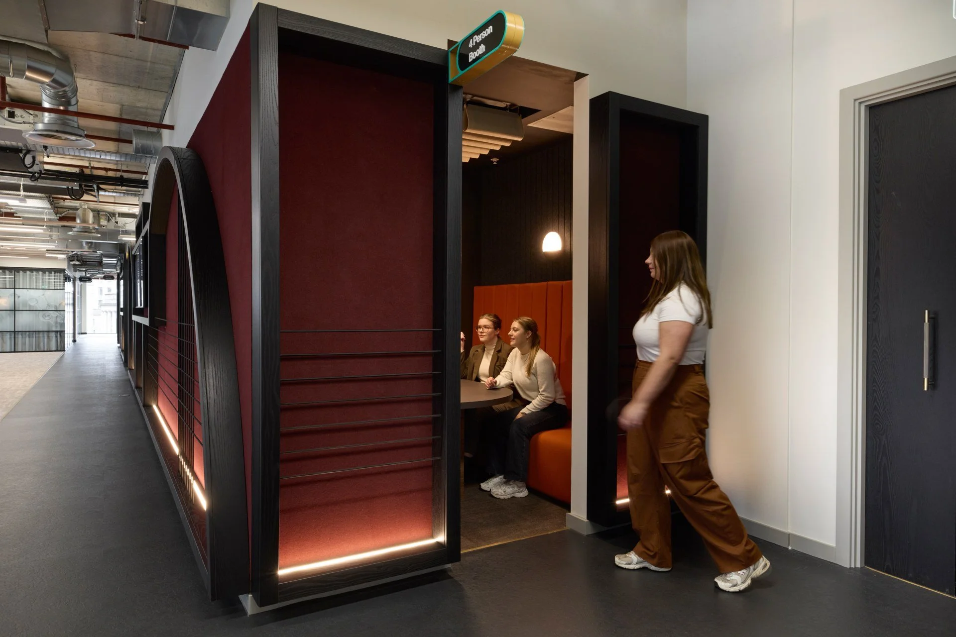 People sitting at a booth labeled '4 Person Booth' in a modern office or restaurant space, with a woman standing nearby.