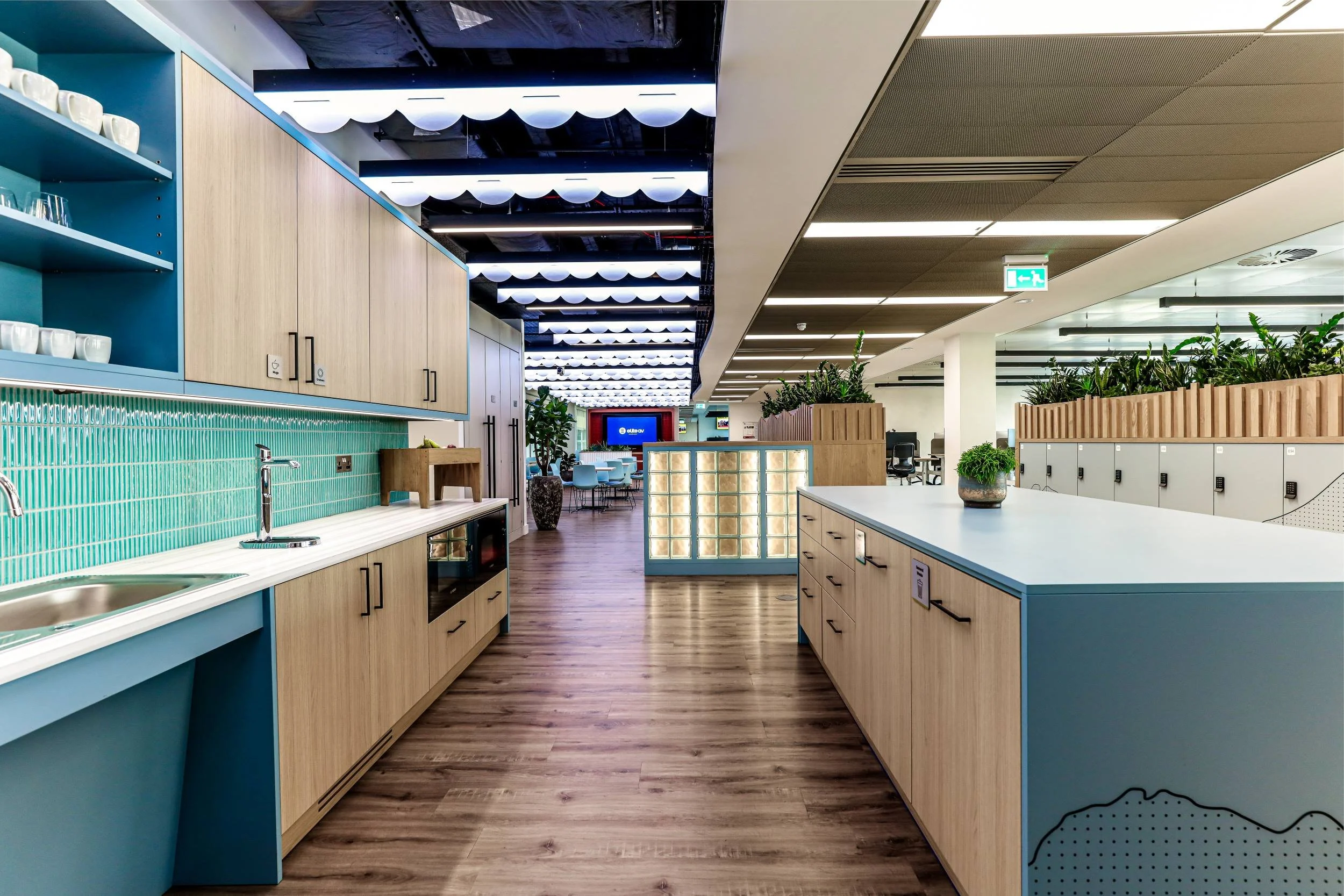Modern office kitchen area with wooden and blue cabinetry, green tiled backsplash, and a built-in sink. In the background, there is a lounge area with seating and a large LED screen on the wall. The space is decorated with potted plants and features a wooden floor and ceiling panels.
