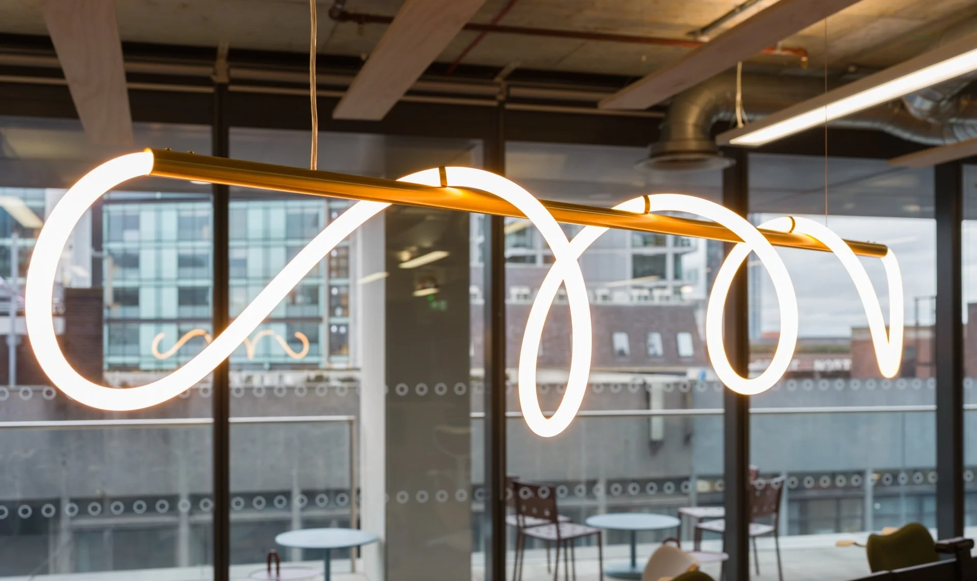 Modern interior with a ceiling light fixture that has a continuous white LED loop design, hanging from a gold rod in front of large floor-to-ceiling windows showing city buildings outside.