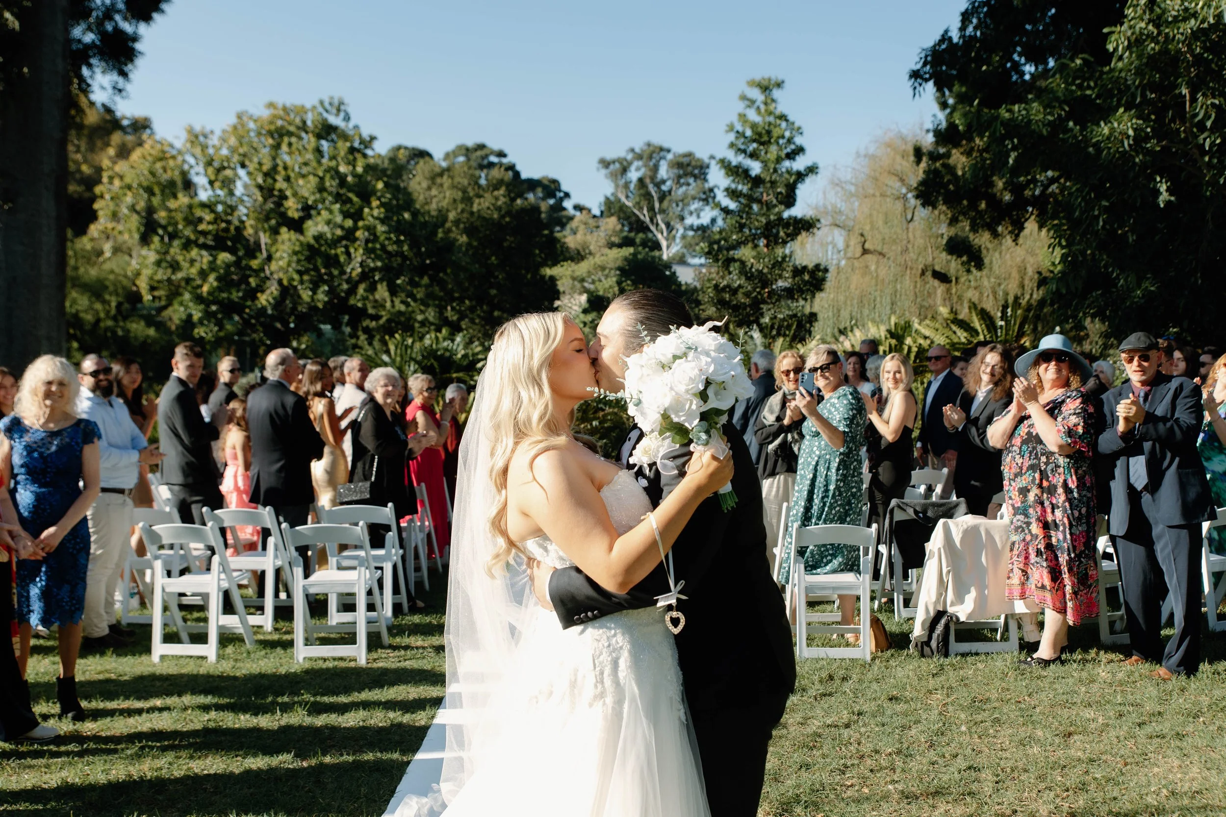 A bride and groom kiss during their outdoor wedding ceremony, with guests standing and sitting in chairs behind them, surrounded by trees and greenery.