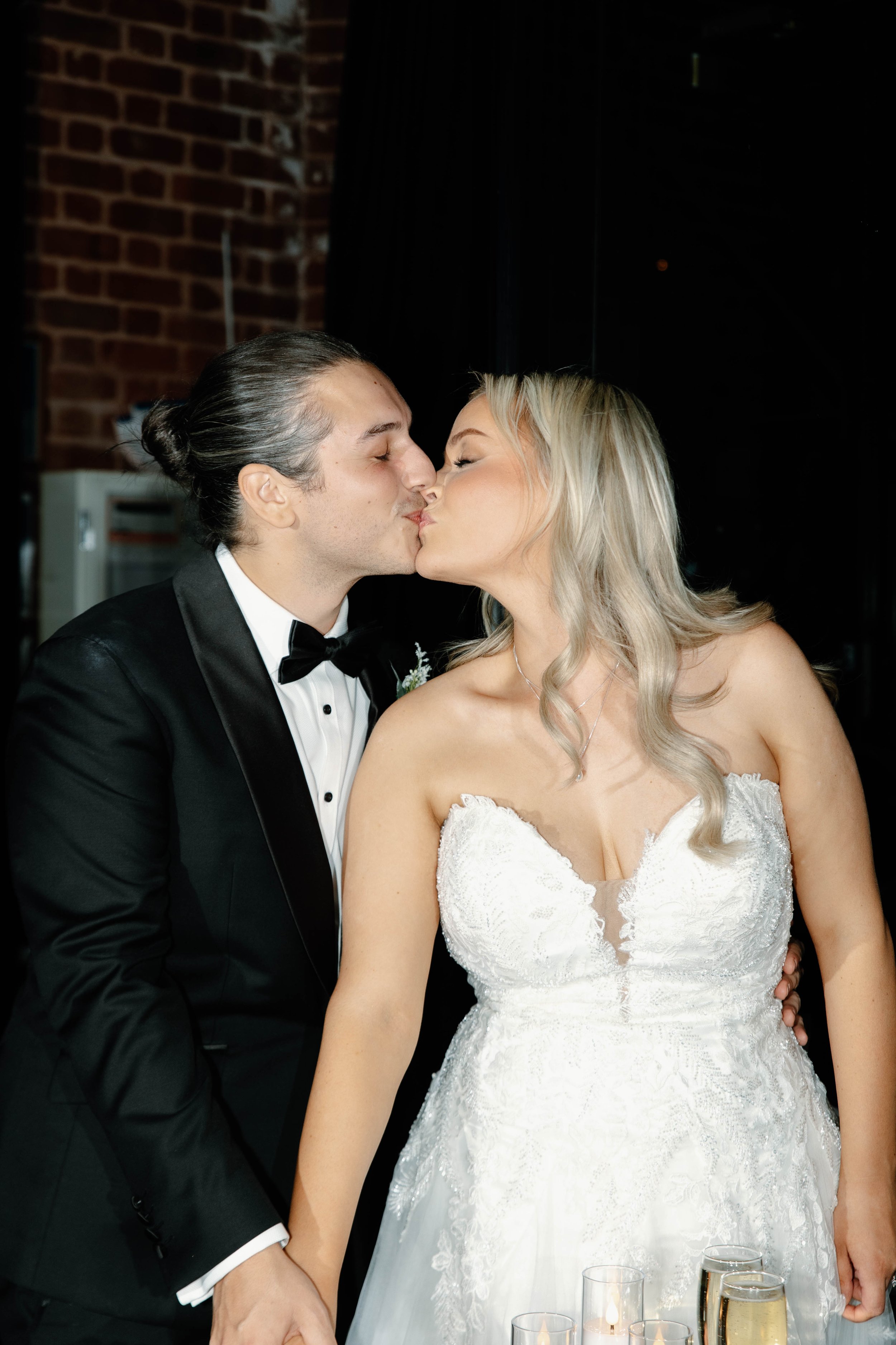 A bride and groom kiss during their wedding reception, with the groom wearing a black tuxedo and the bride in a white lace wedding dress, holding hands.