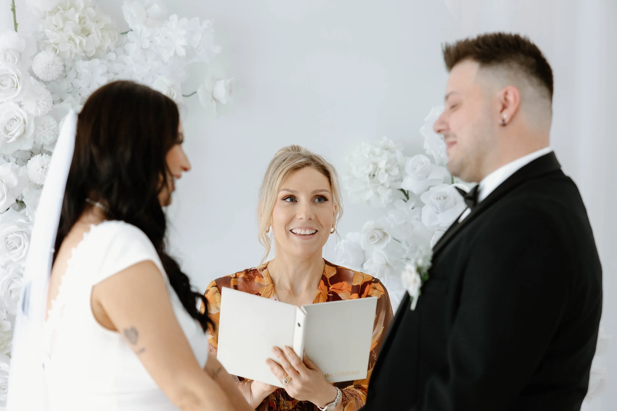 A woman officiant conducting a wedding ceremony, with a bride and groom standing before her in front of a white floral backdrop.