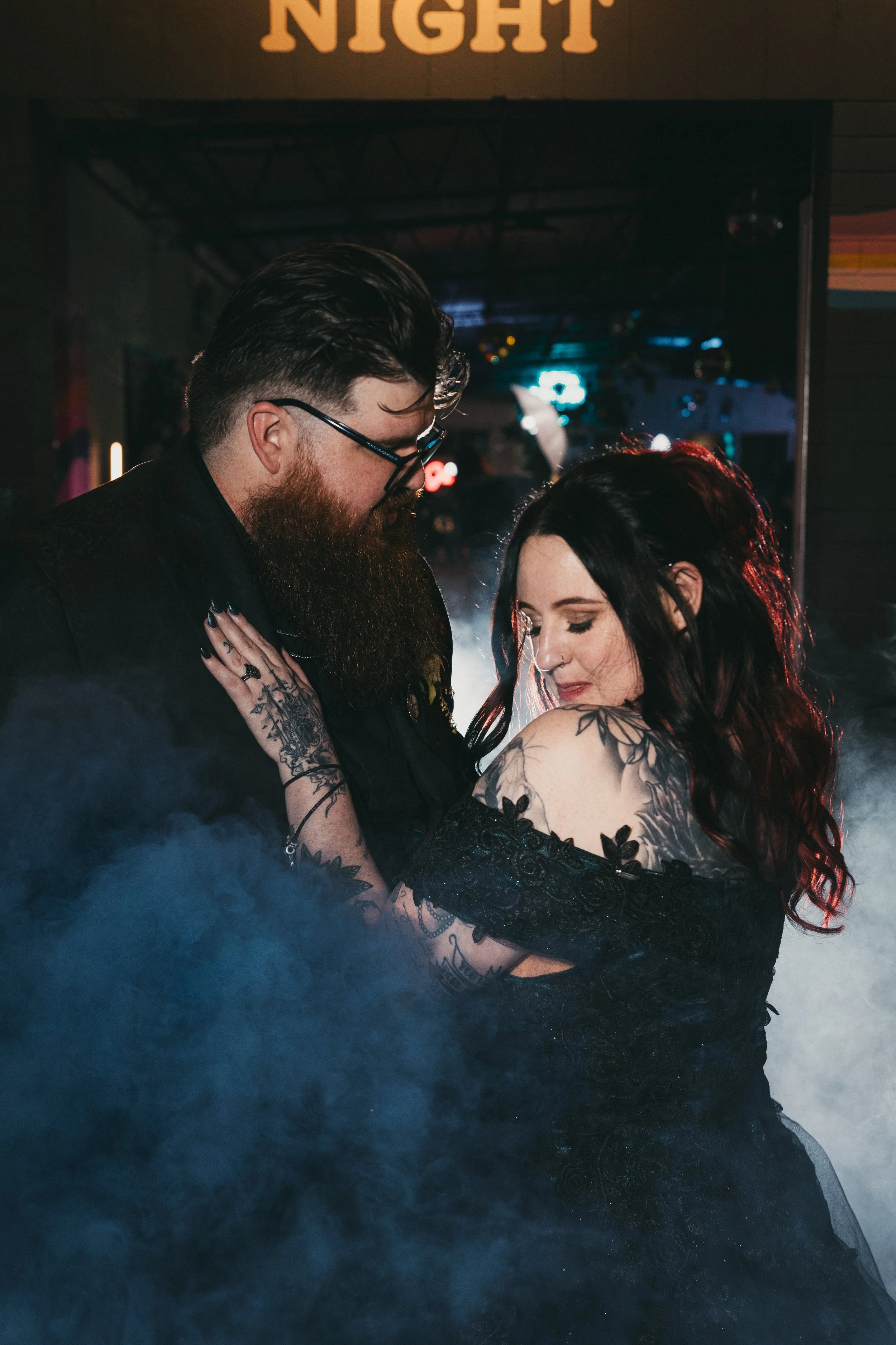 A man and woman dancing closely together in a dimly lit environment with neon lights, with the word 'NIGHT' visible on a sign overhead.