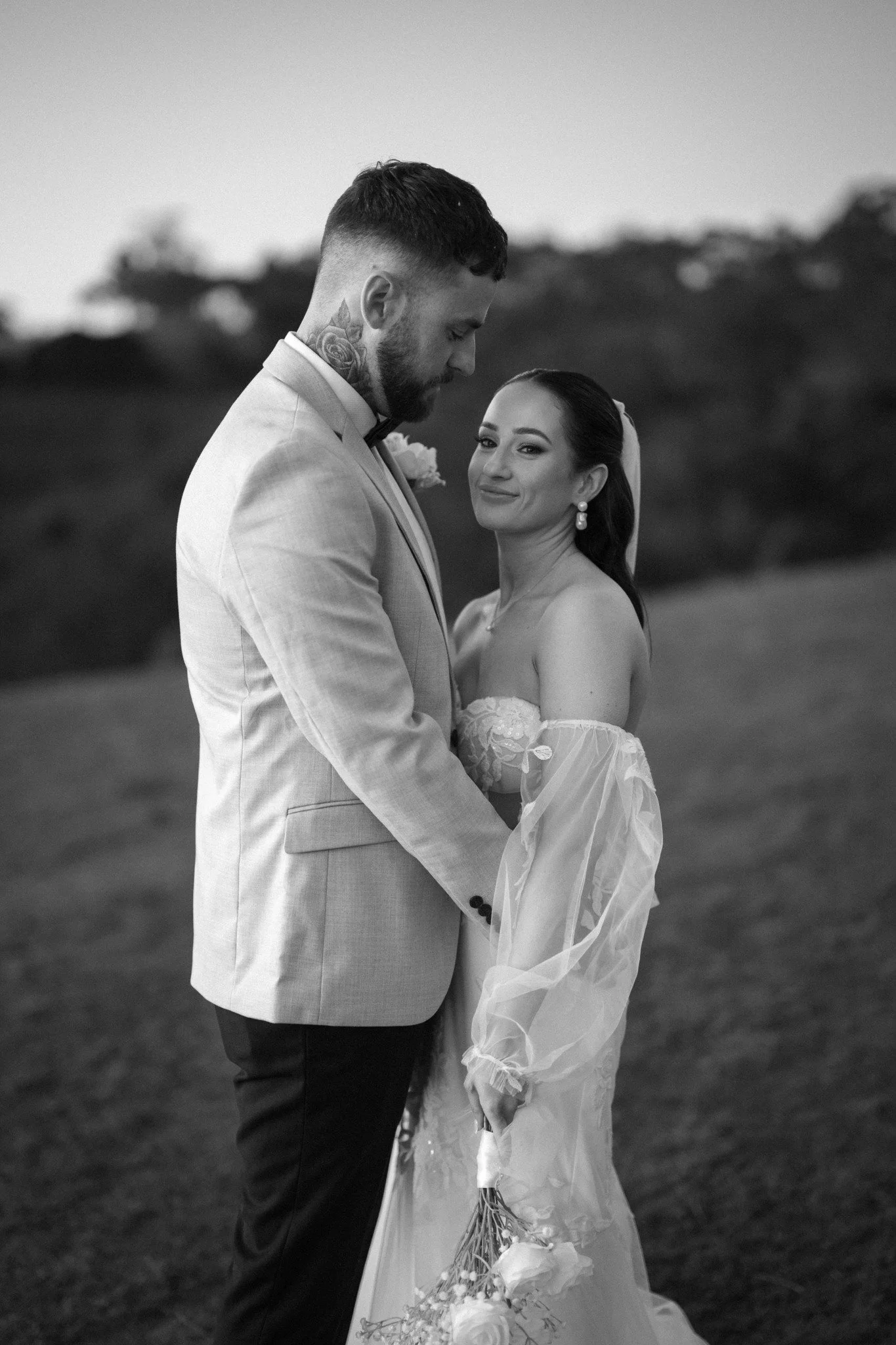 Black and white photo of a bride and groom standing outdoors, holding hands, with the groom looking down and the bride smiling at the camera, holding a bouquet of flowers.