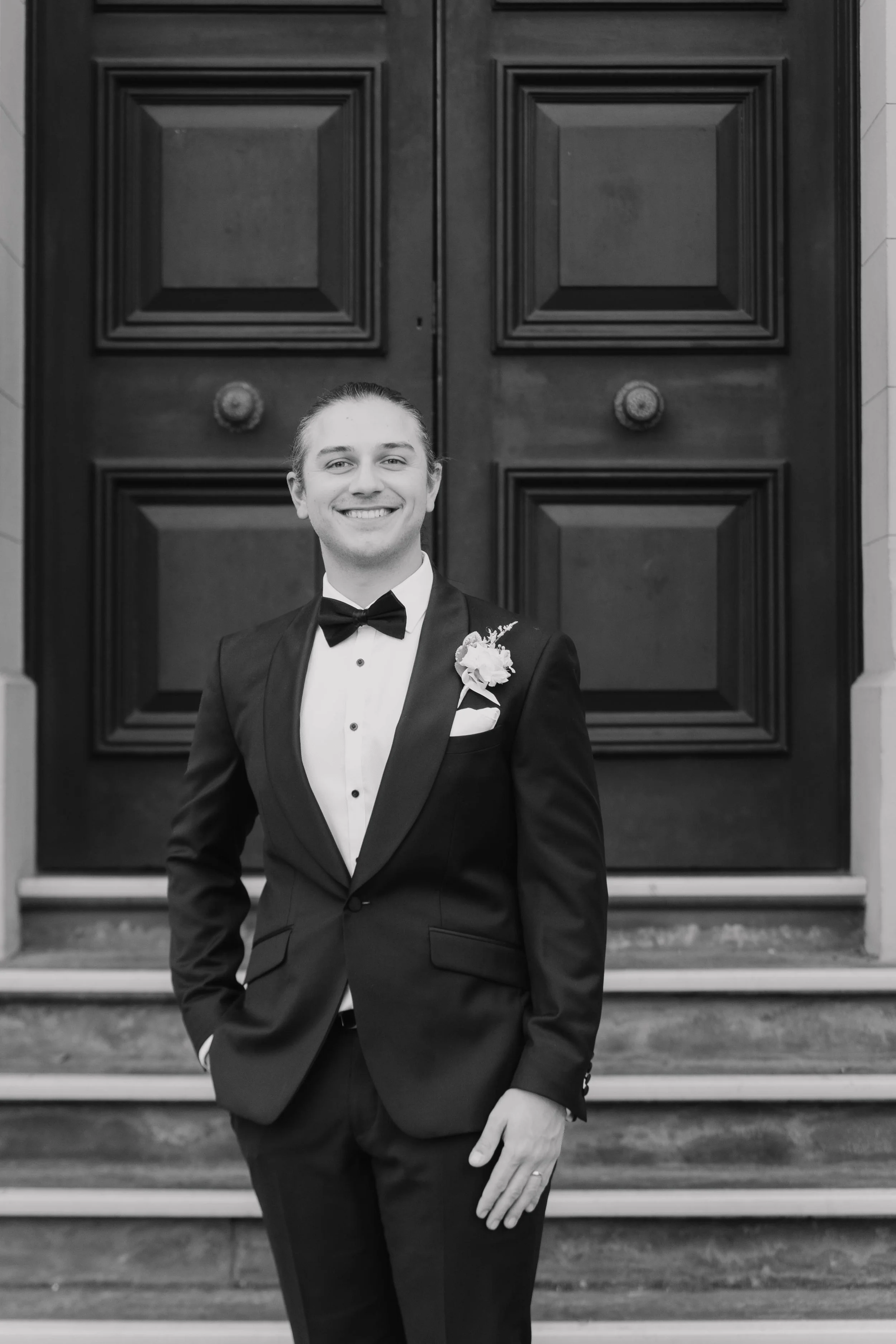 A young man in a tuxedo with a bow tie and boutonnière, smiling and standing on the steps in front of a large, decorative wooden door.
