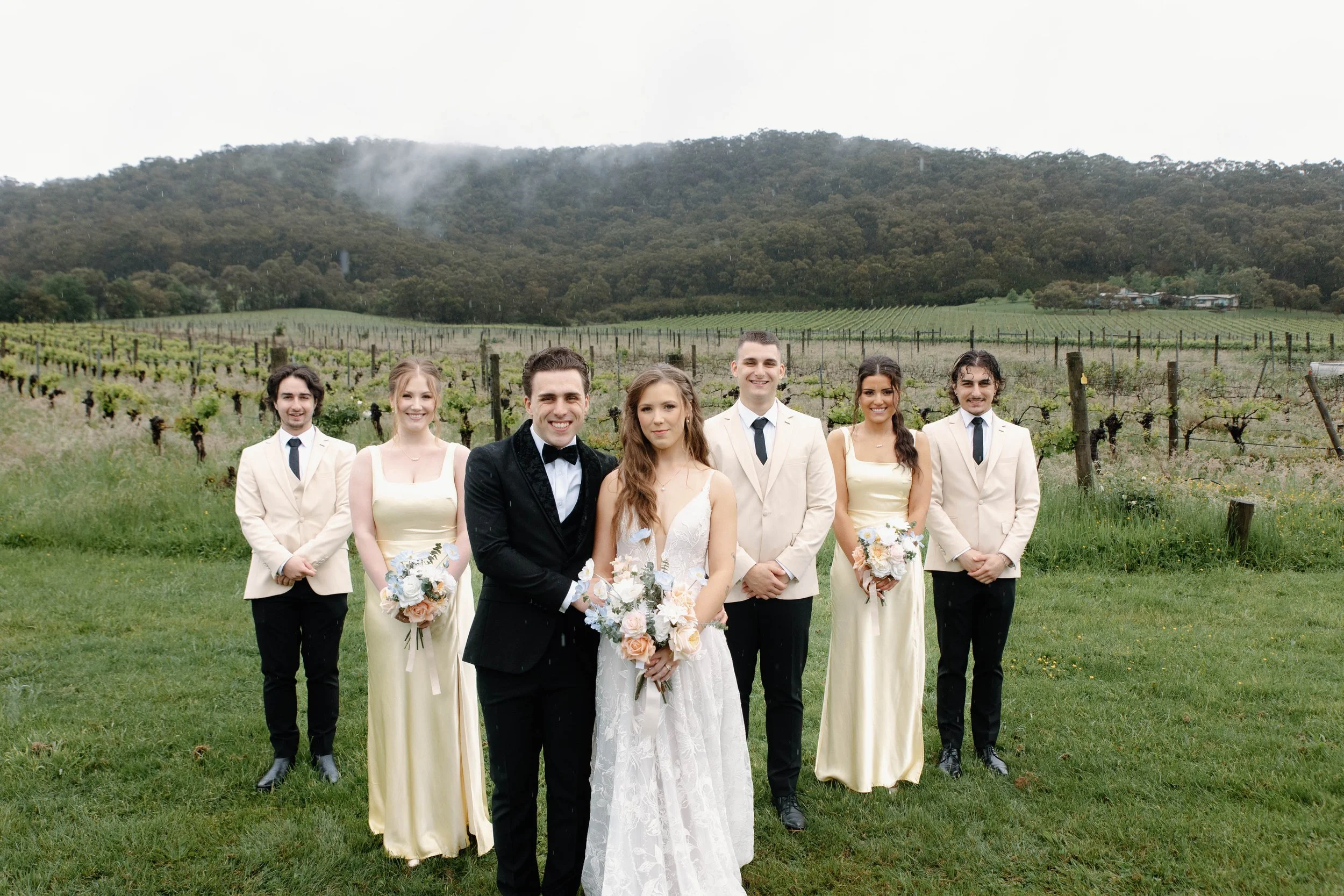 A wedding party of seven people standing outdoors on green grass with a vineyard and mountains in the background. The bride and groom are in the center, with the bride in a white wedding dress holding a bouquet, and the groom in a black tuxedo. Four 